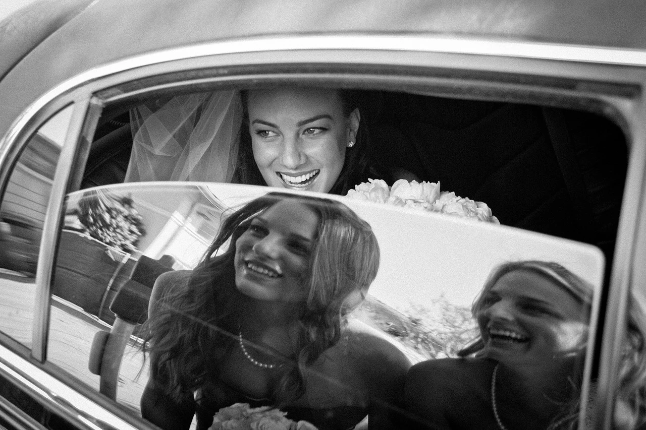 A bride smiles from inside a car while women outside appear reflected in the glass.