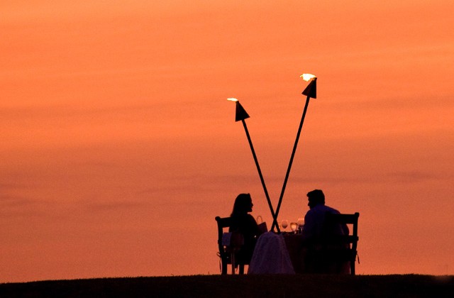Two people sit at a table between flaming torches against an orange sunset sky.