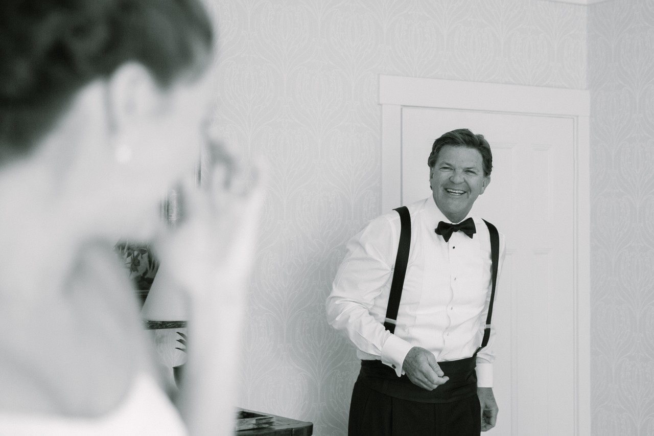 A groom smiles broadly toward a bride seen in the foreground in black and white.