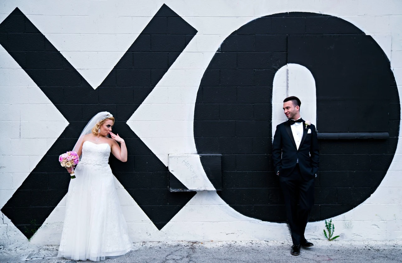 A bride and groom stand apart in front of oversized black shapes painted on a white wall.