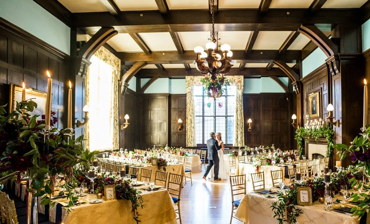 A decorated reception room features wooden ceiling beams, chandeliers and arranged dining tables.