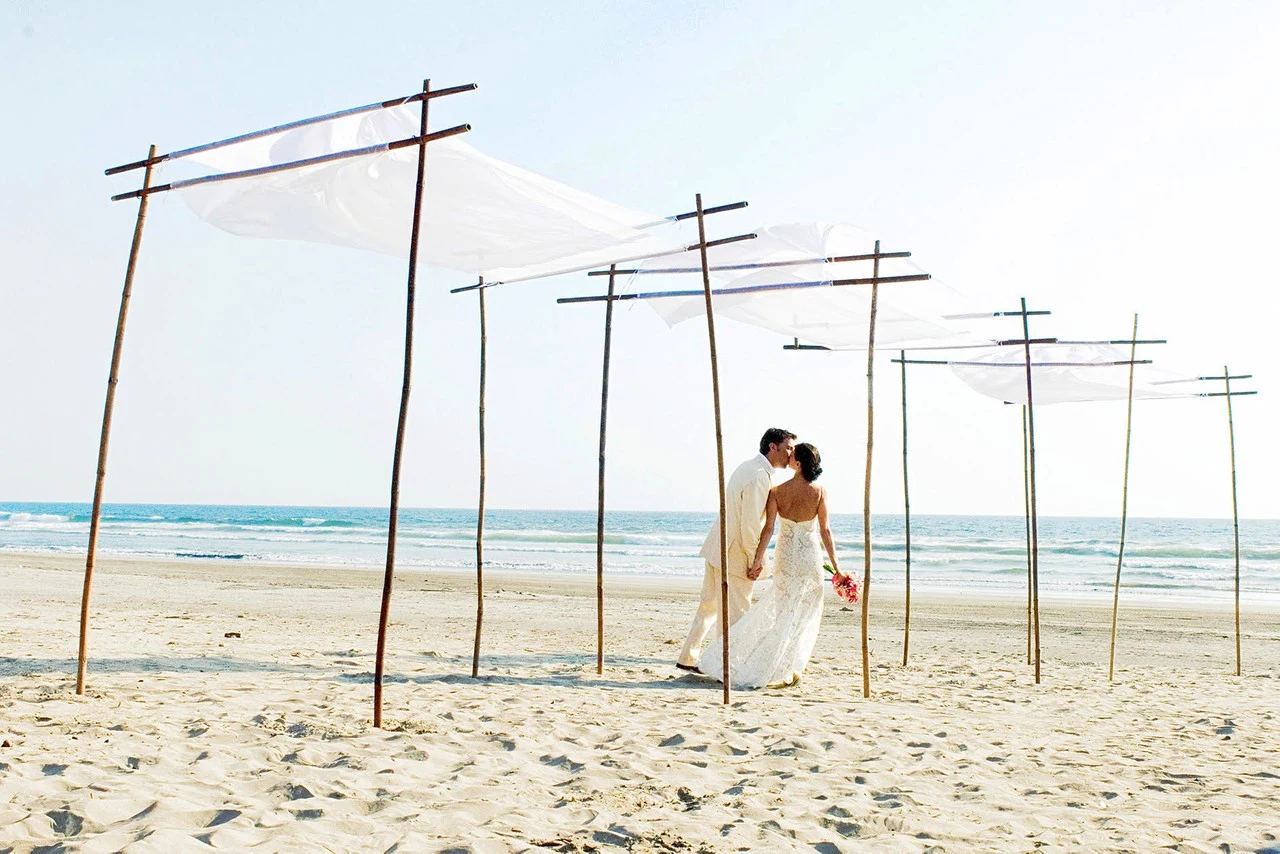 A bride walks barefoot under simple poles and floating white fabric on a sandy beach.