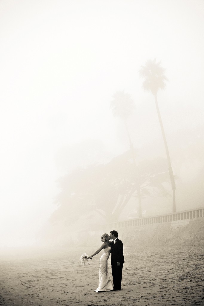 A couple embraces on a foggy beach with a faint palm tree behind them in monochrome.