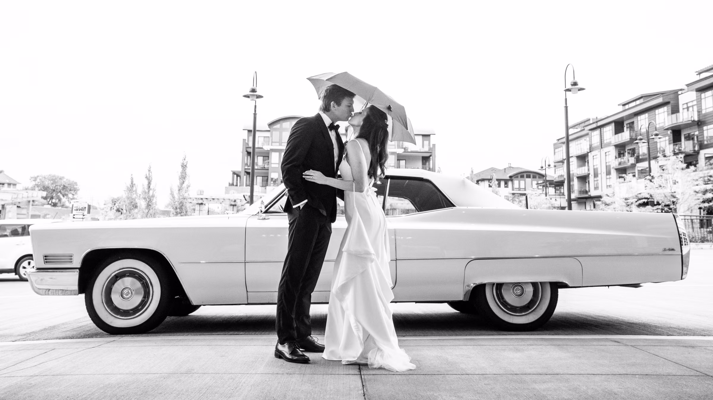 Ali and Drew kiss under a clear umbrella beside the white vintage Cadillac