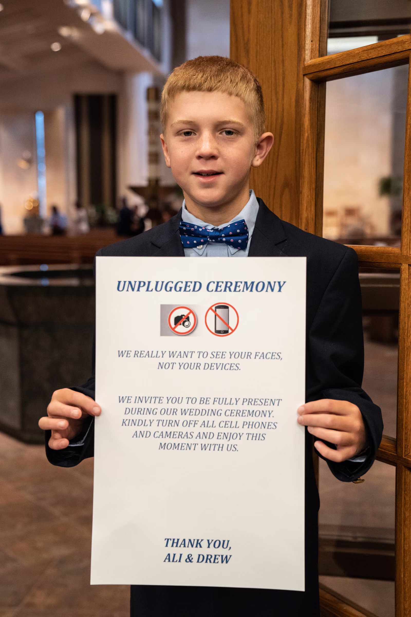 A young ring bearer holds Ali and Drew’s unplugged ceremony sign inside the church