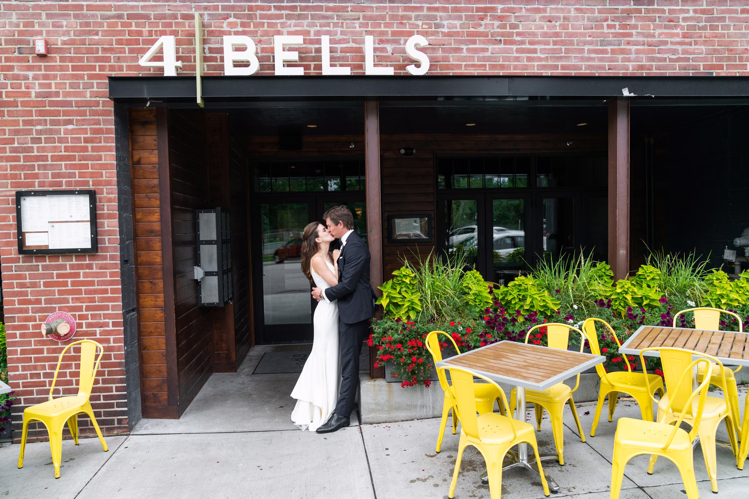 Ali and Drew kiss outside the brick entrance of 4 Bells beside bright yellow patio chairs
