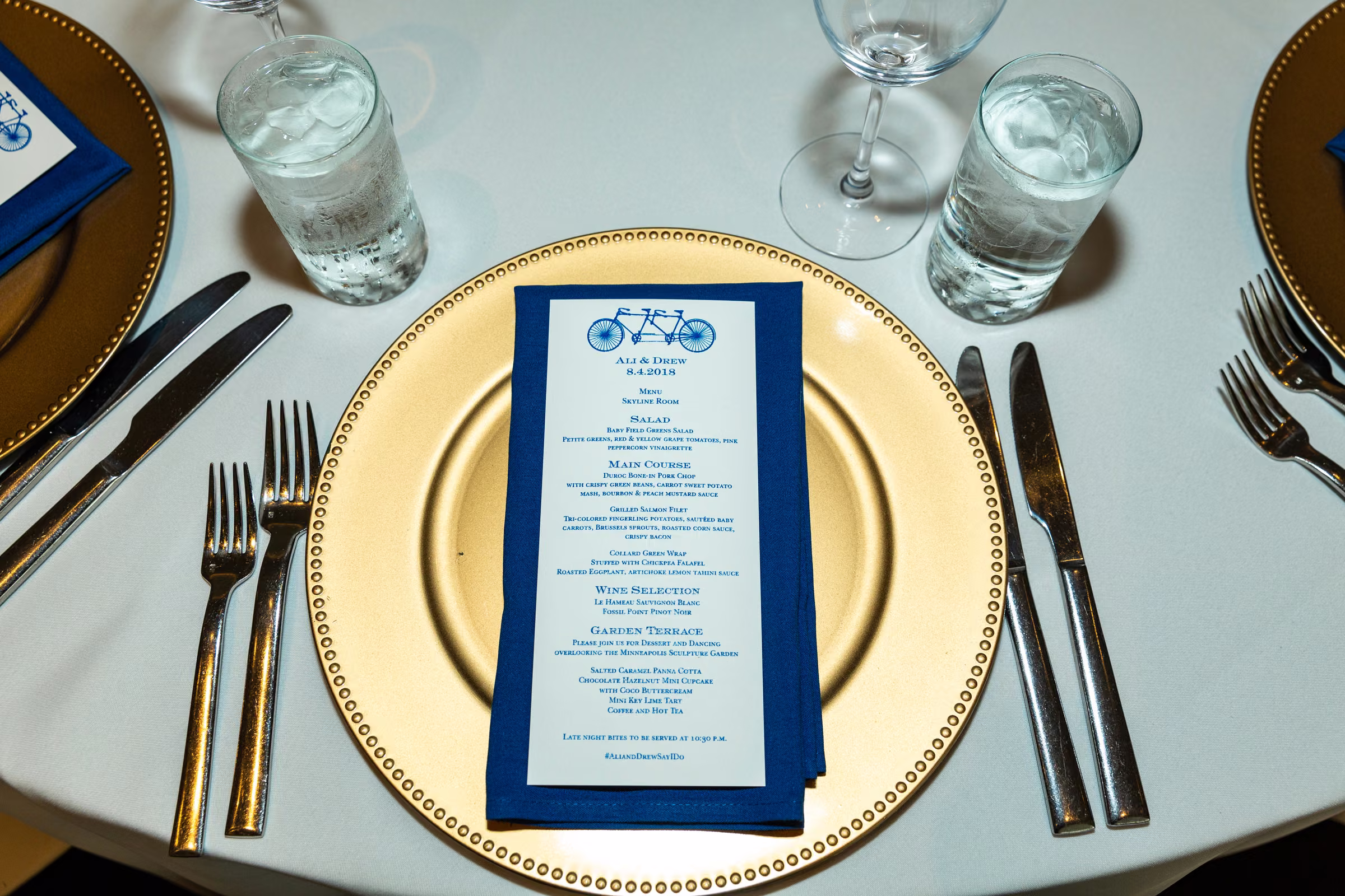 A blue-bordered wedding menu rests on a gold charger beside neatly arranged silverware and glasses