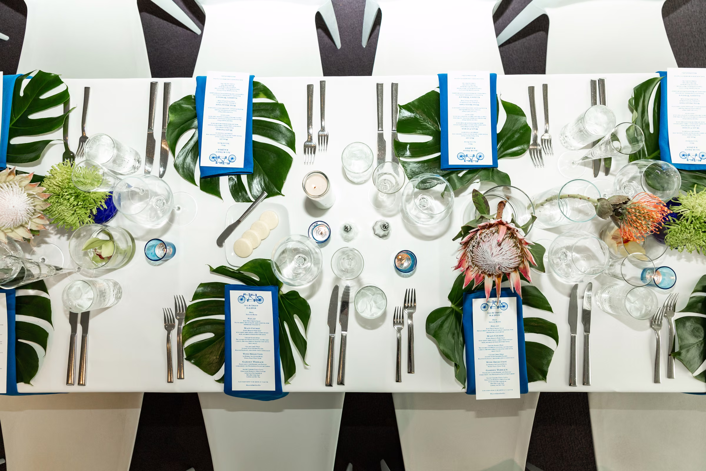 Overhead view of wedding menus, tropical leaves, flowers, glassware, and gold chargers on the reception table