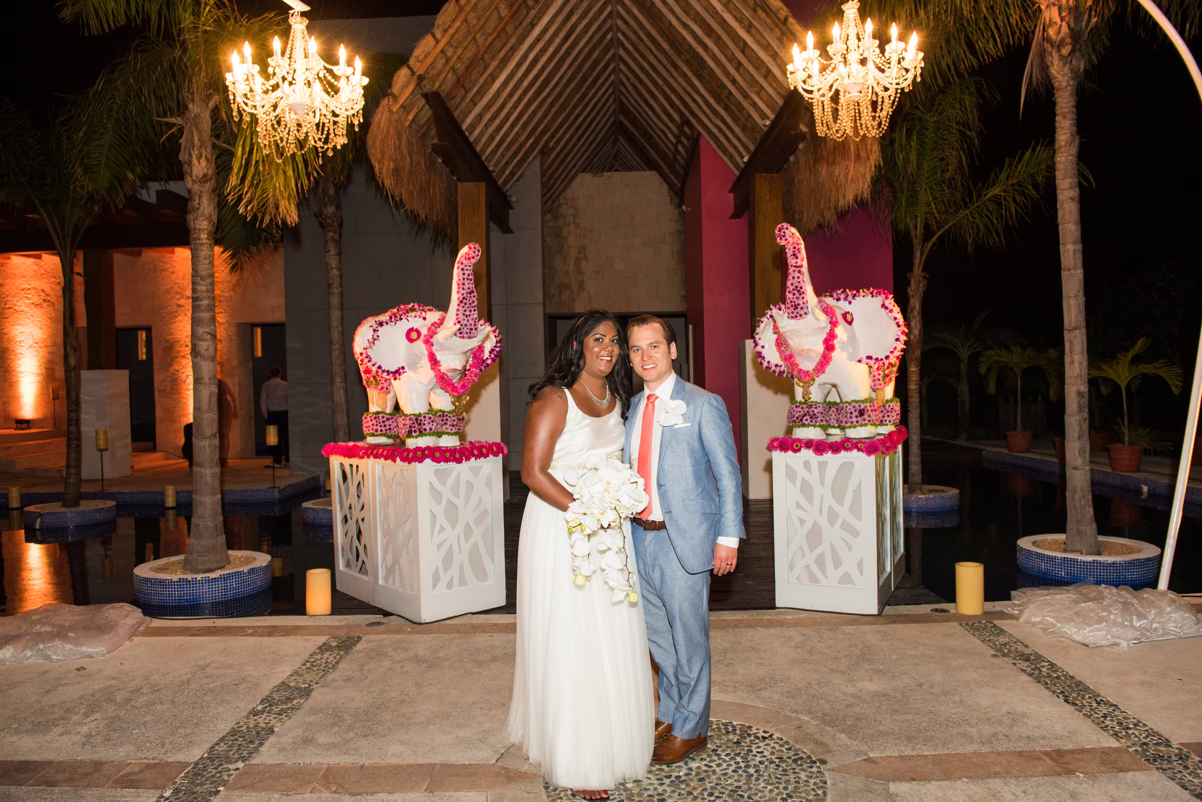 Avanti and Tony stand at an illuminated reception entrance beneath chandeliers and beside floral displays