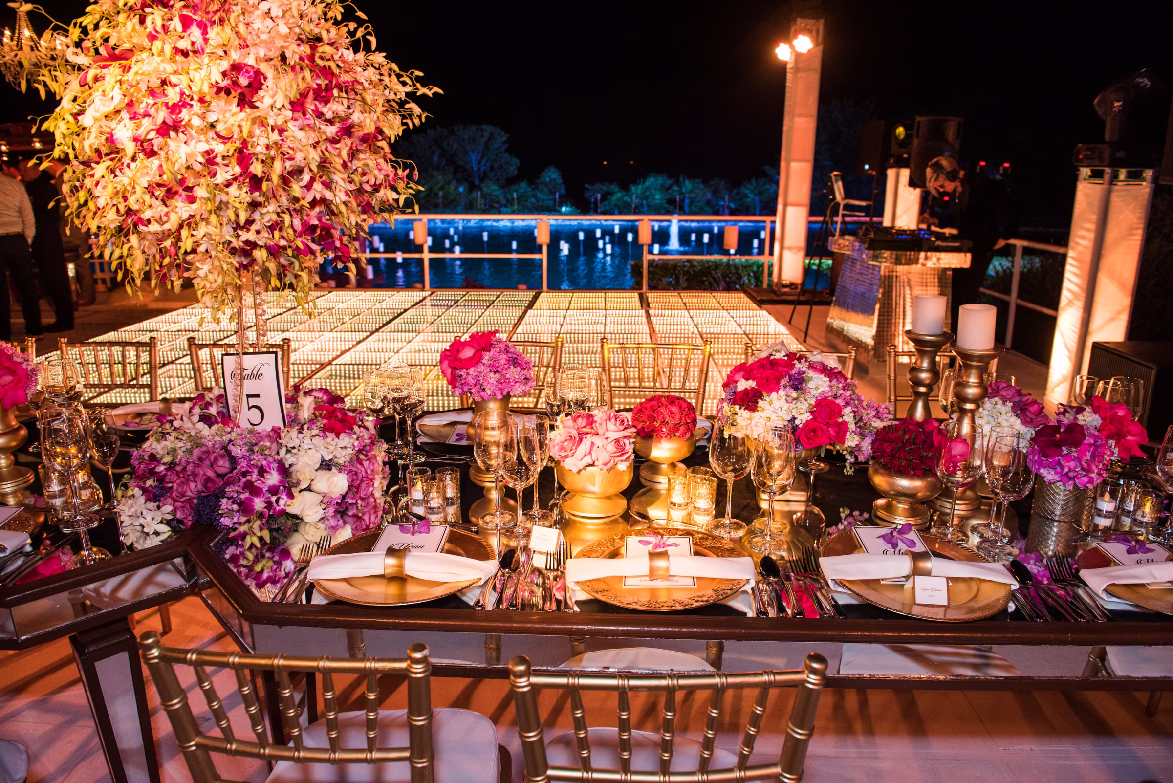A reception table with flowers, candles, and glassware is arranged beneath warm lights