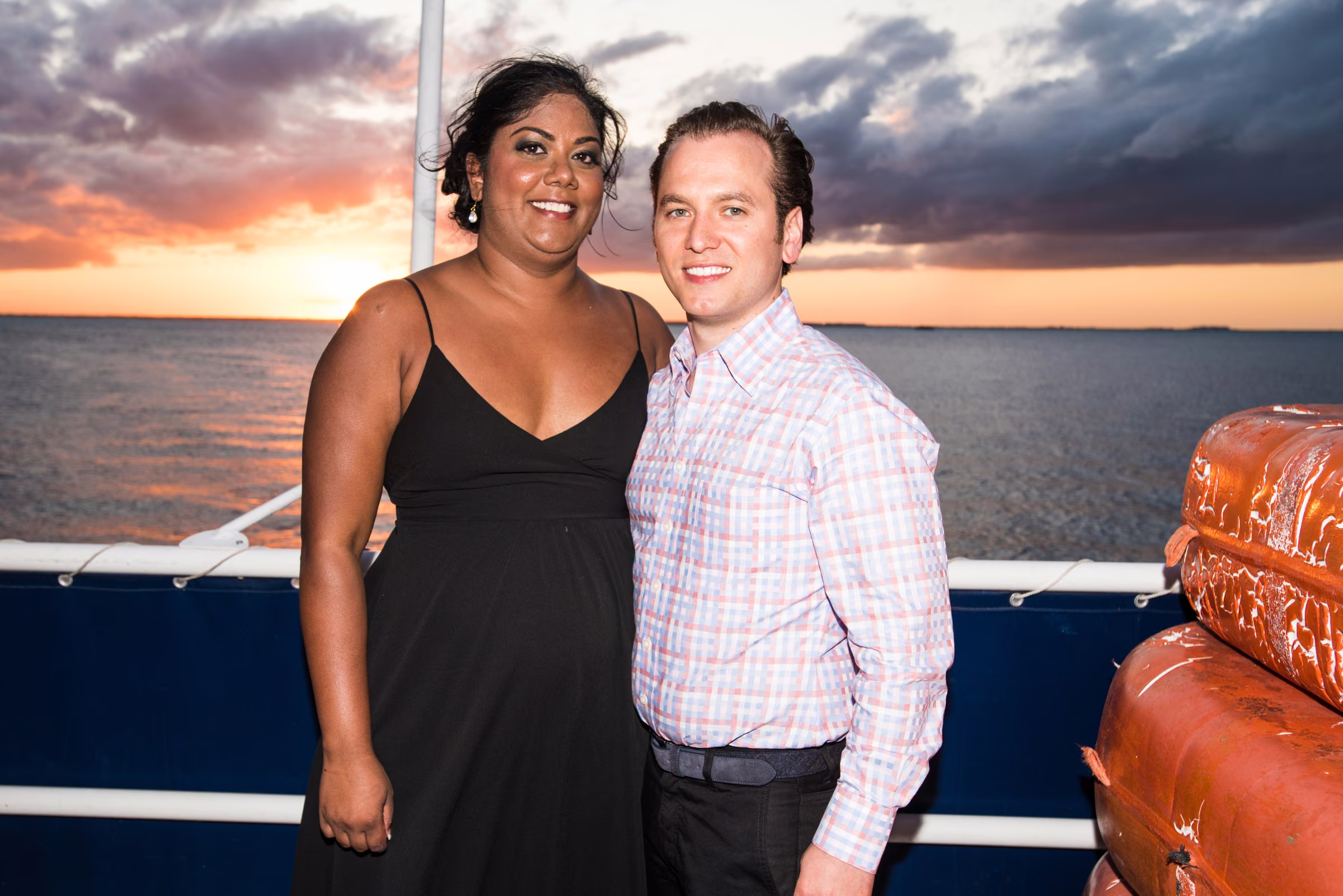 Avanti and Tony smile together on the boat with a colorful sunset over the water behind them