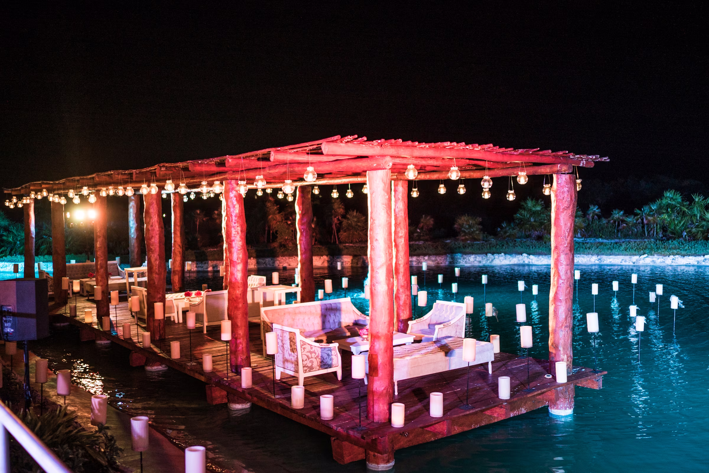An illuminated dining pavilion sits beside the water with candles along its edges