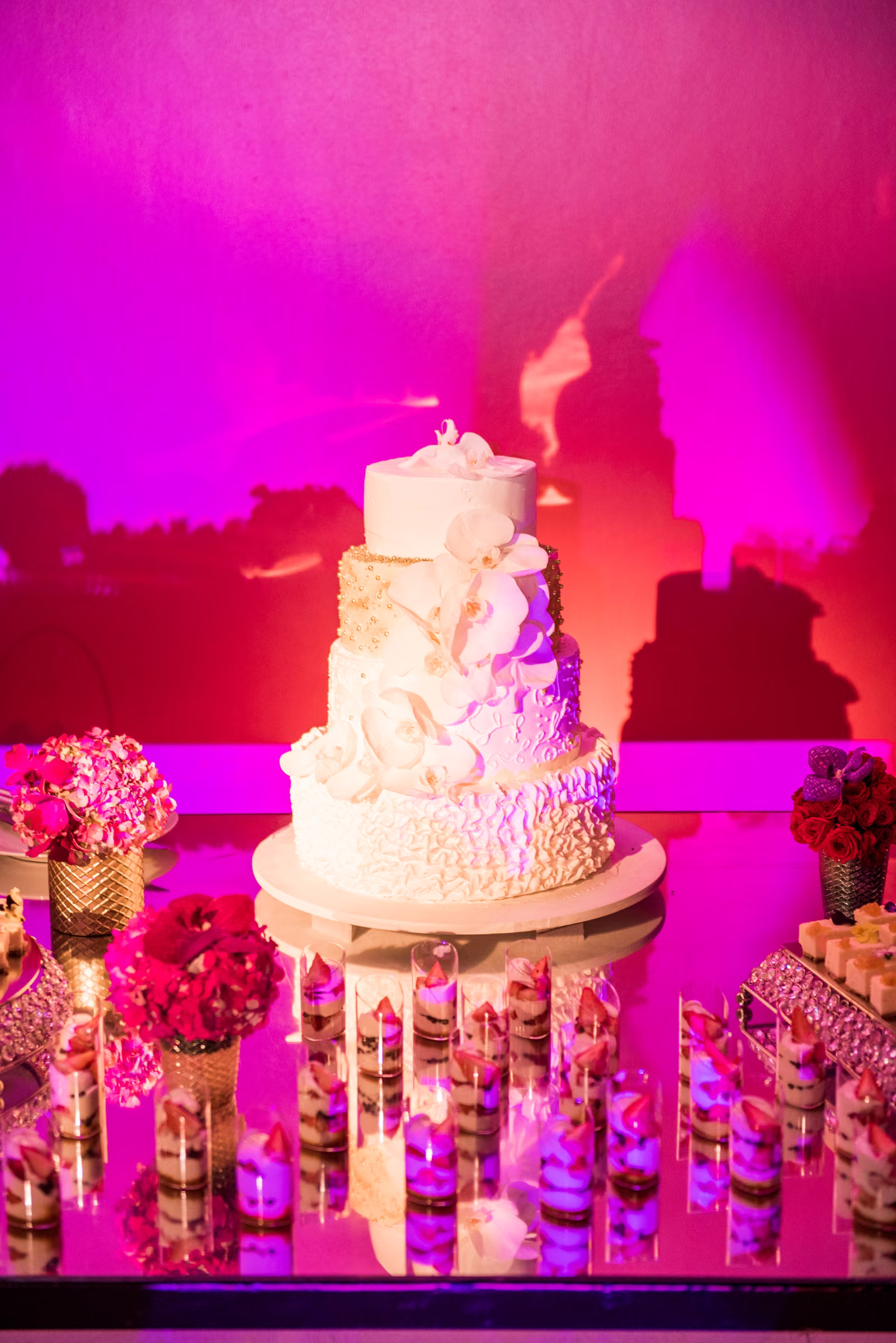 A tiered white wedding cake with floral details stands among flowers and candles under pink lighting