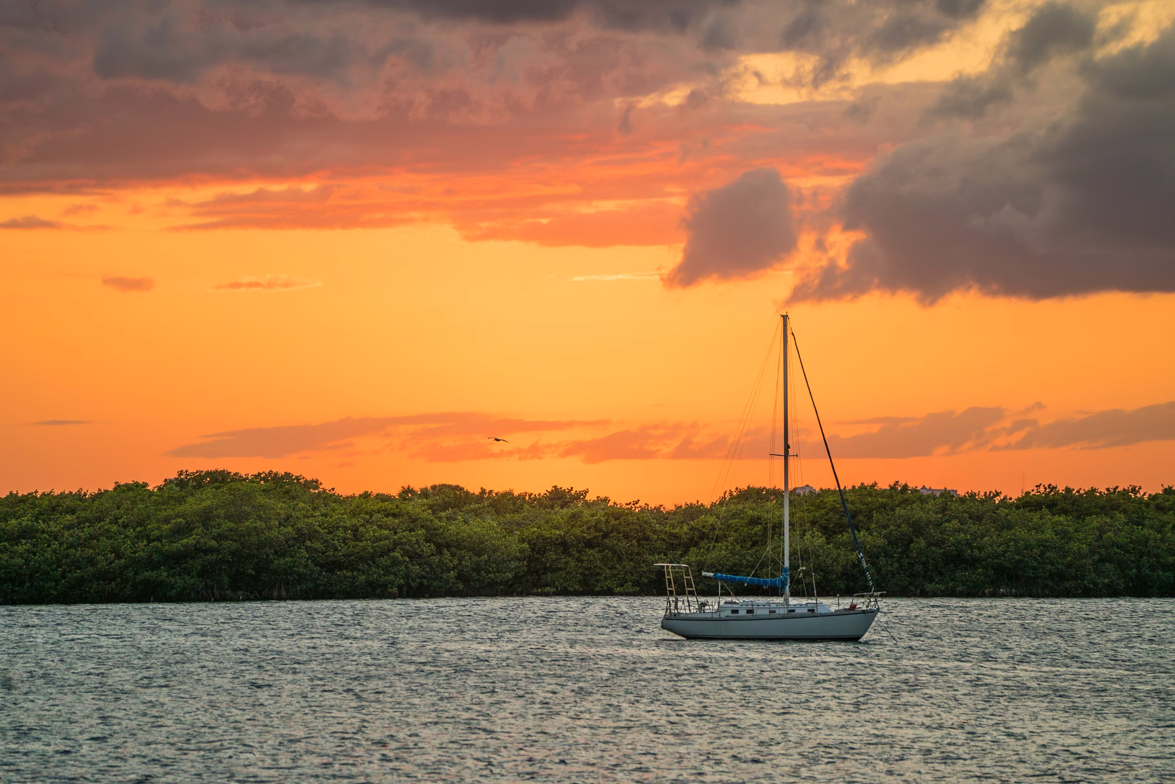 An orange sunset lights the clouds above a sailboat near the shore