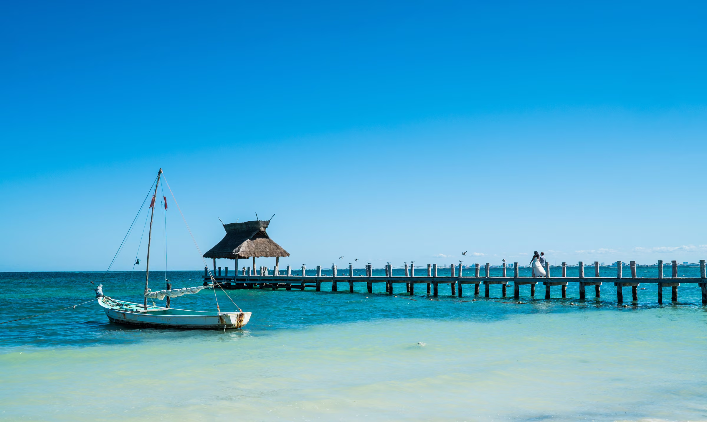 Avanti and Tony appear small on a long pier across bright turquoise water beside a sailboat