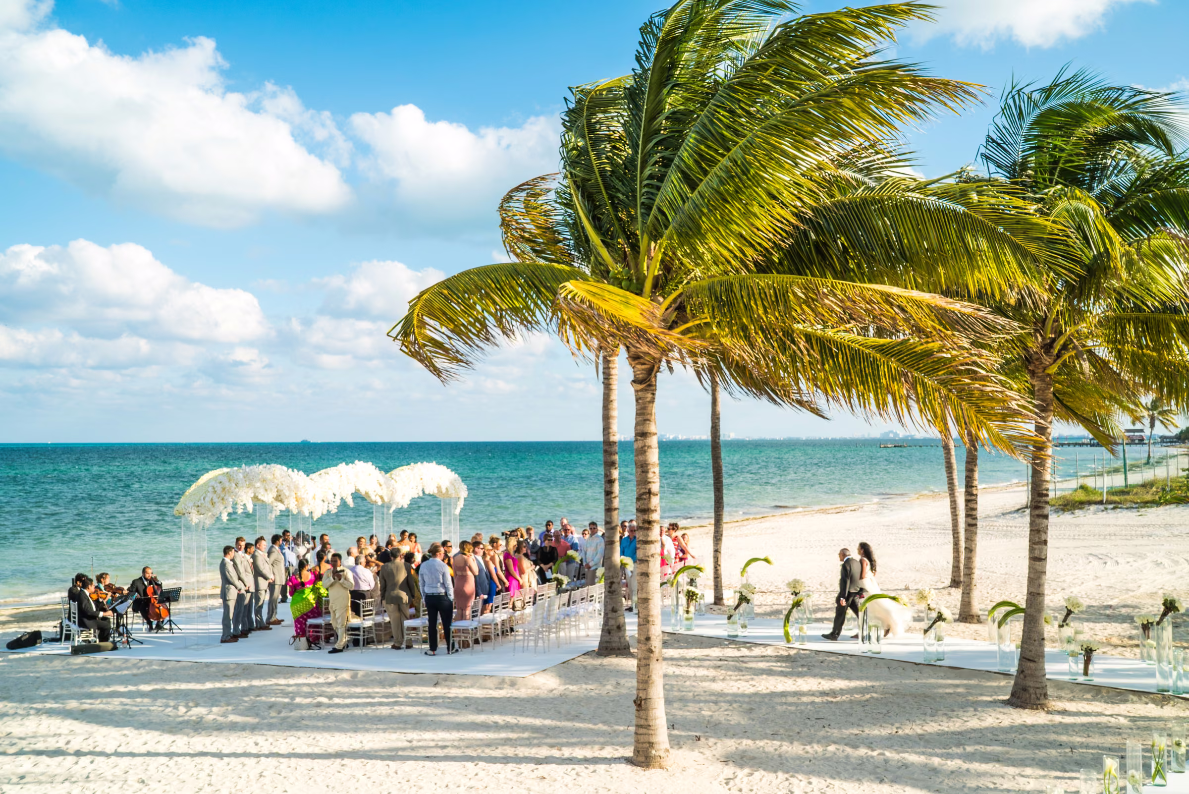 An expansive beach view shows Avanti walking with her father toward the ceremony beneath palms