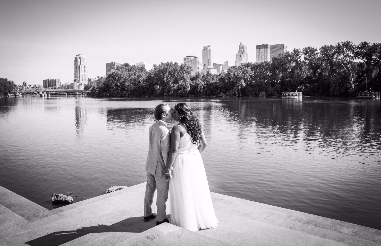 A black and white photograph of Avanti and Tony kissing beside the Mississippi River with downtown Minneapolis behind them