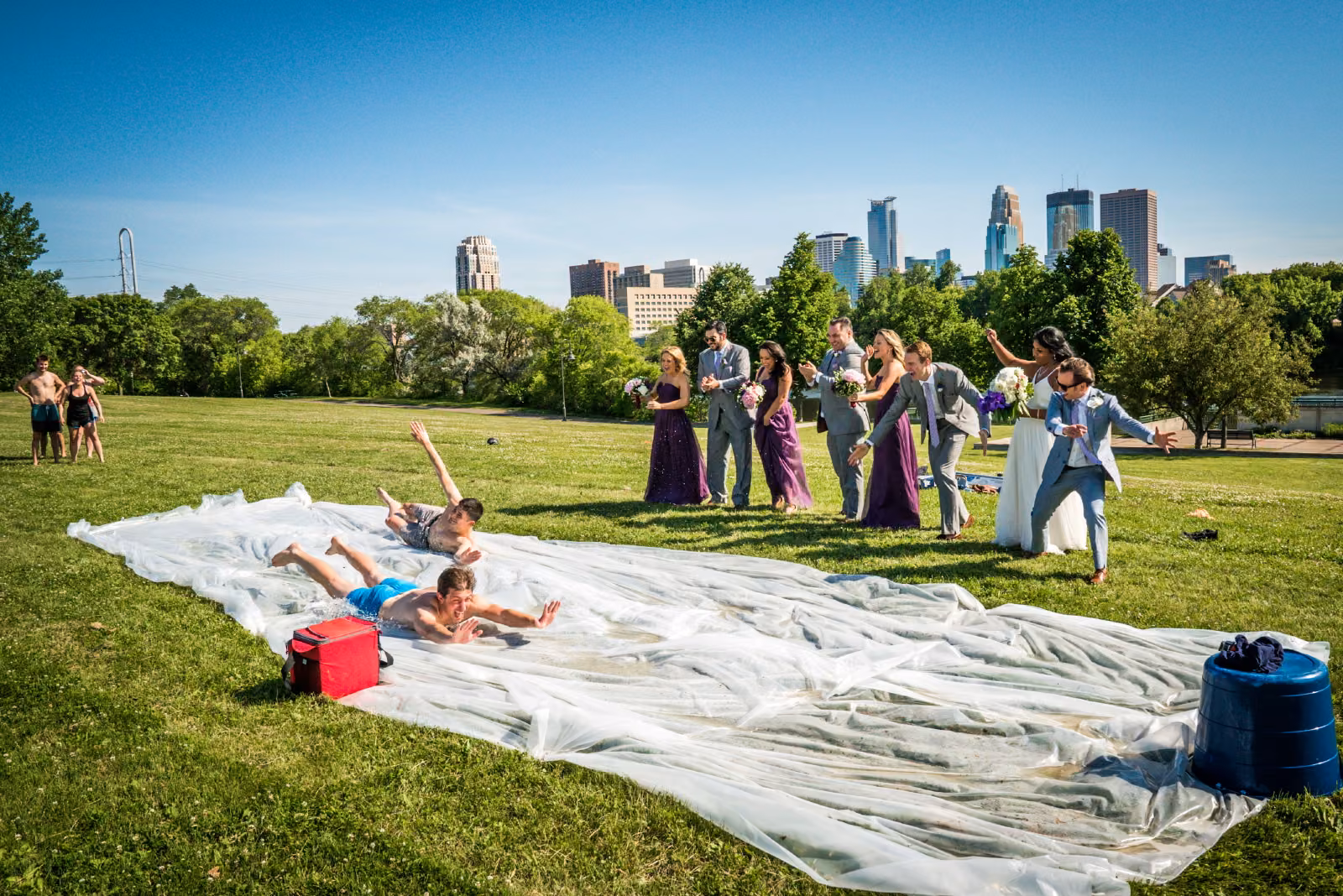 Wedding guests in purple dresses and gray suits cheer beside a homemade slip-and-slide on a Minneapolis park lawn with the skyline behind them