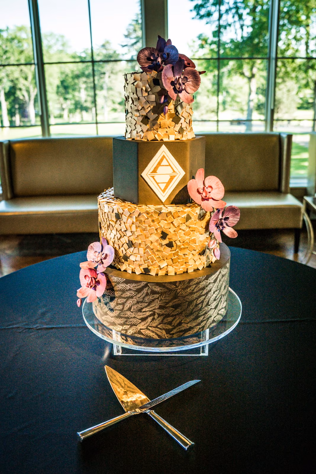 A tall wedding cake with textured gold and dark tiers, flowers, and the couple’s geometric monogram at Interlachen Country Club