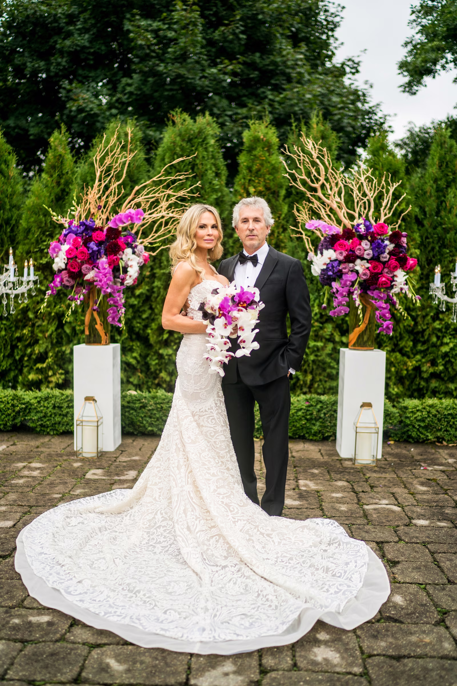 Bill and Daiva stand together between colorful floral arrangements in the backyard