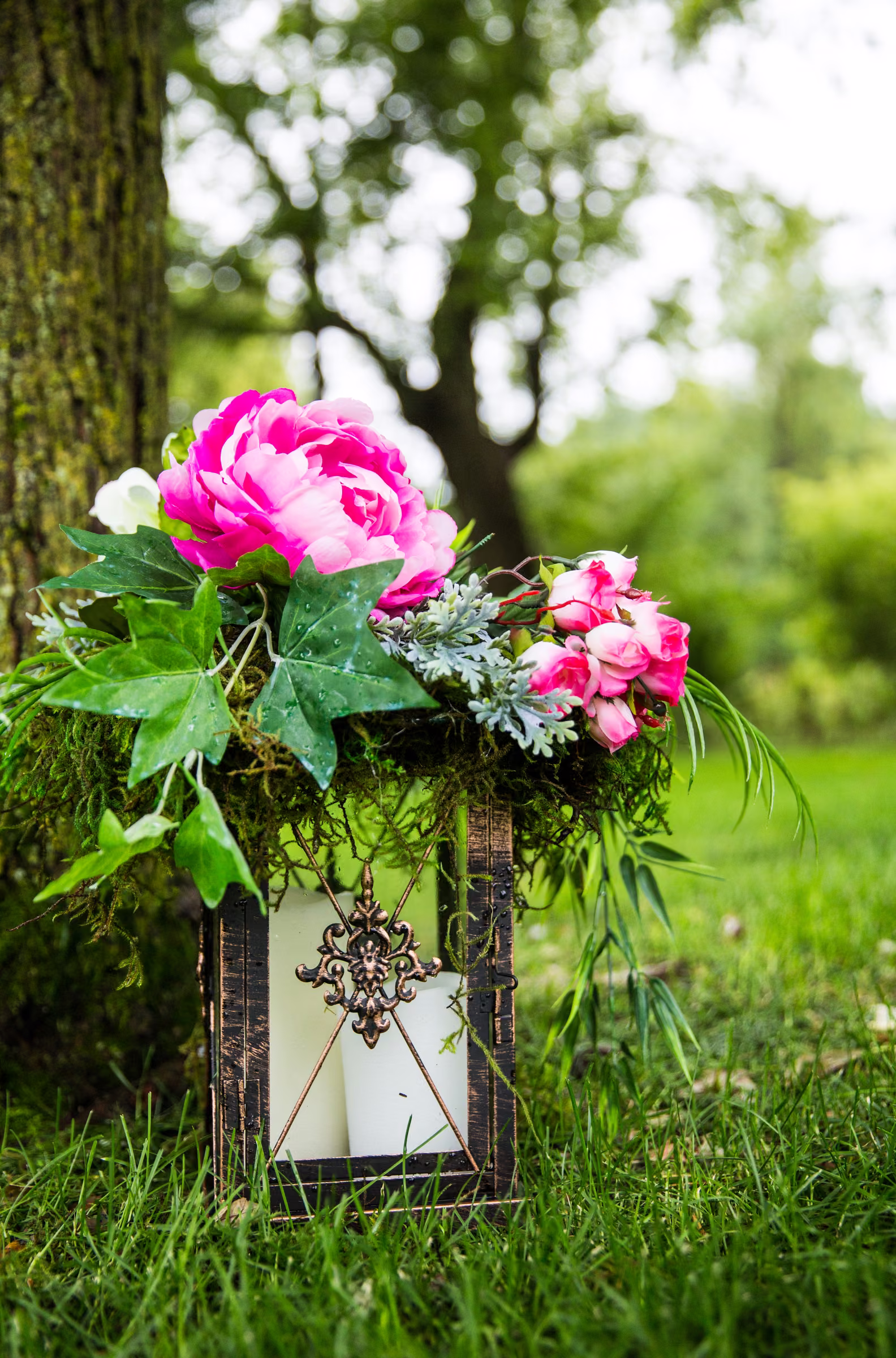 A candle lantern topped with pink flowers sits in the grass beside a tree