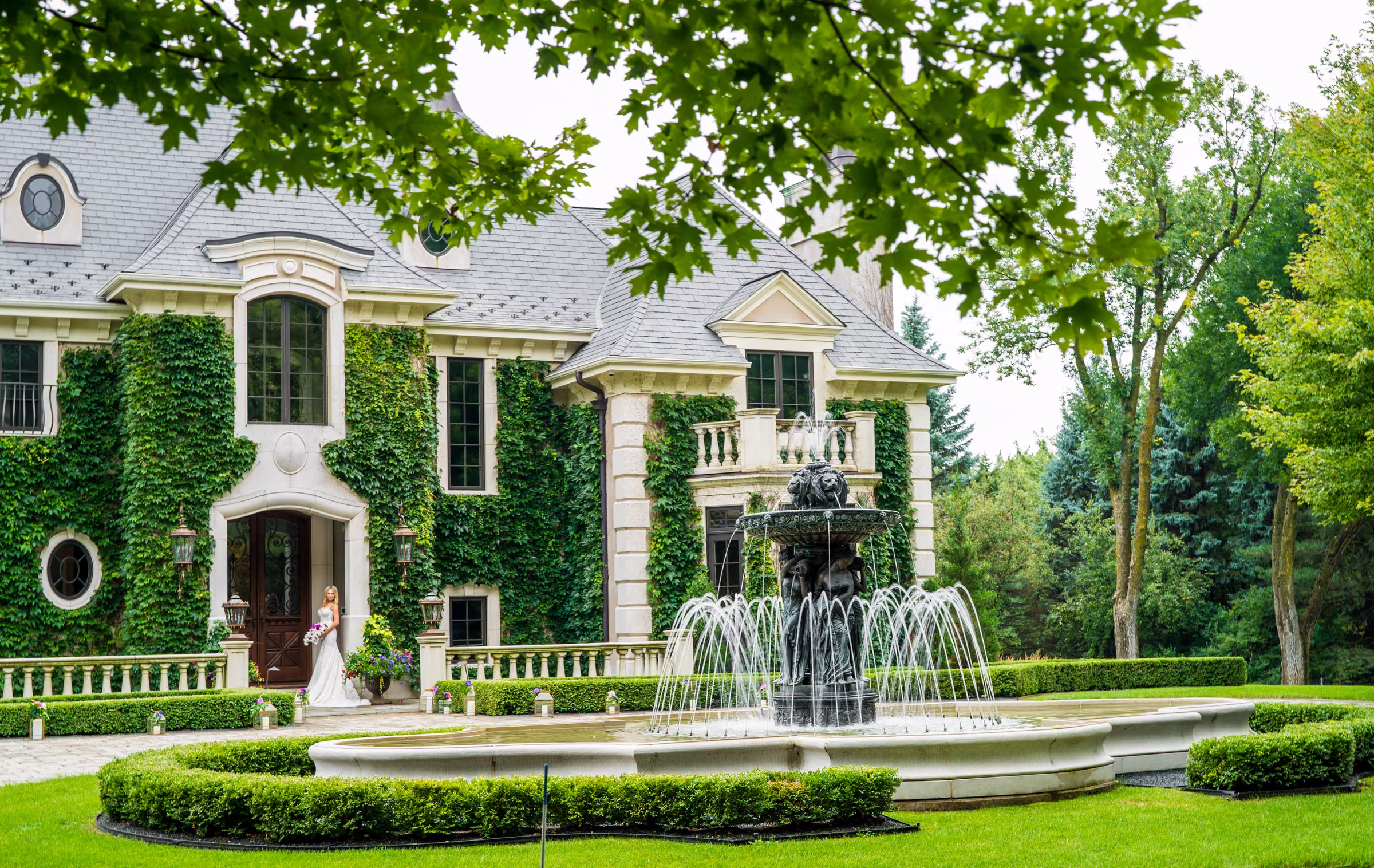 Daiva stands in the front doorway of a large ivy-covered private estate with a fountain in the foreground
