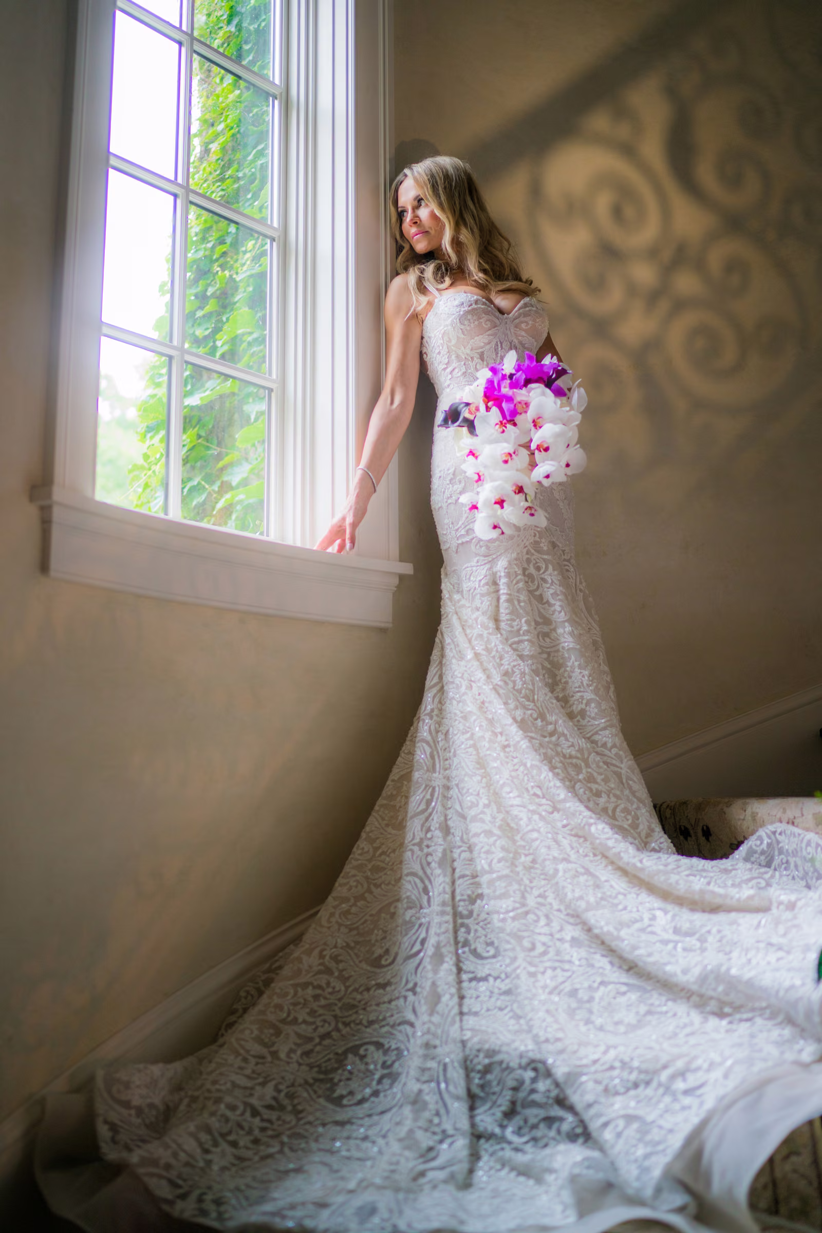 Daiva stands on the staircase beside a tall window with her dress trailing behind her