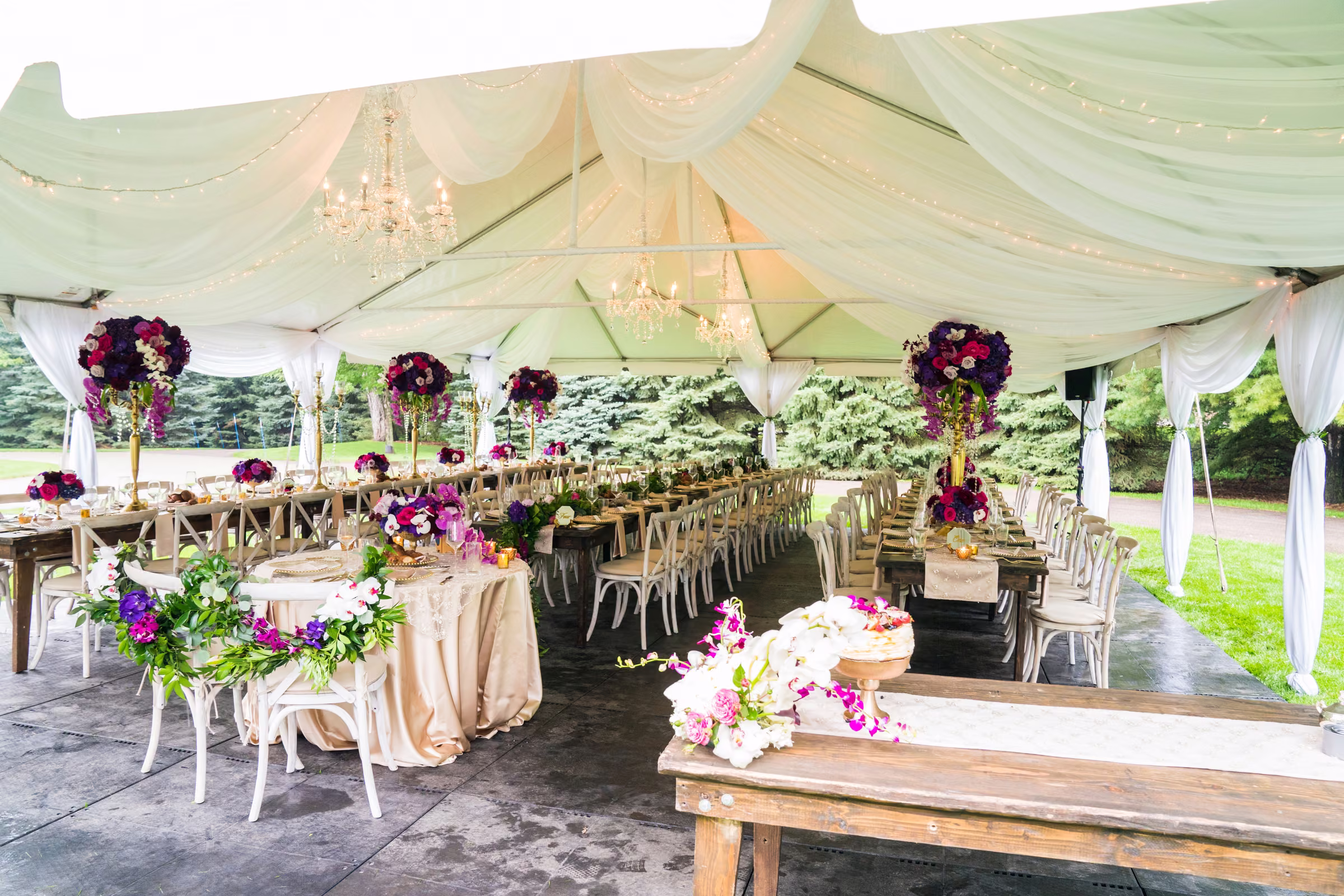A white reception tent is filled with flowers, chandeliers, and long dining tables