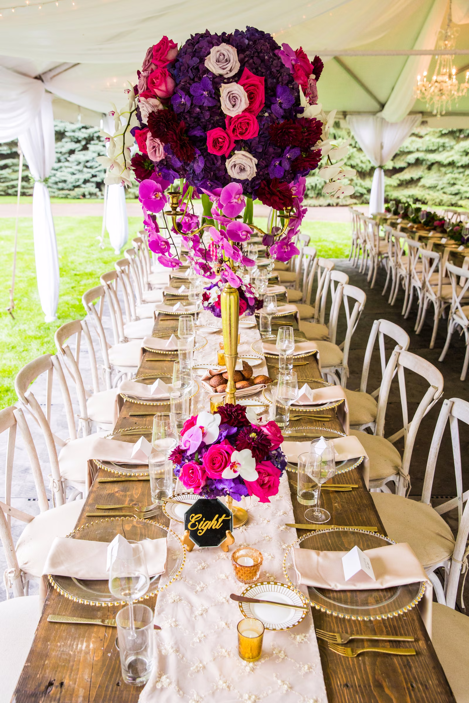 Tall pink and purple floral centerpieces line a long reception table beneath the tent