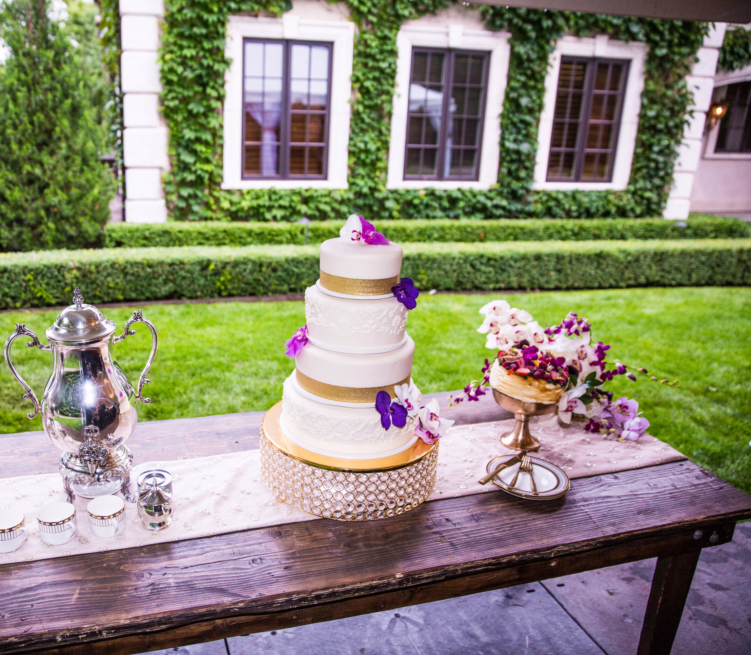 A tiered white wedding cake with gold bands and purple flowers stands outside the estate