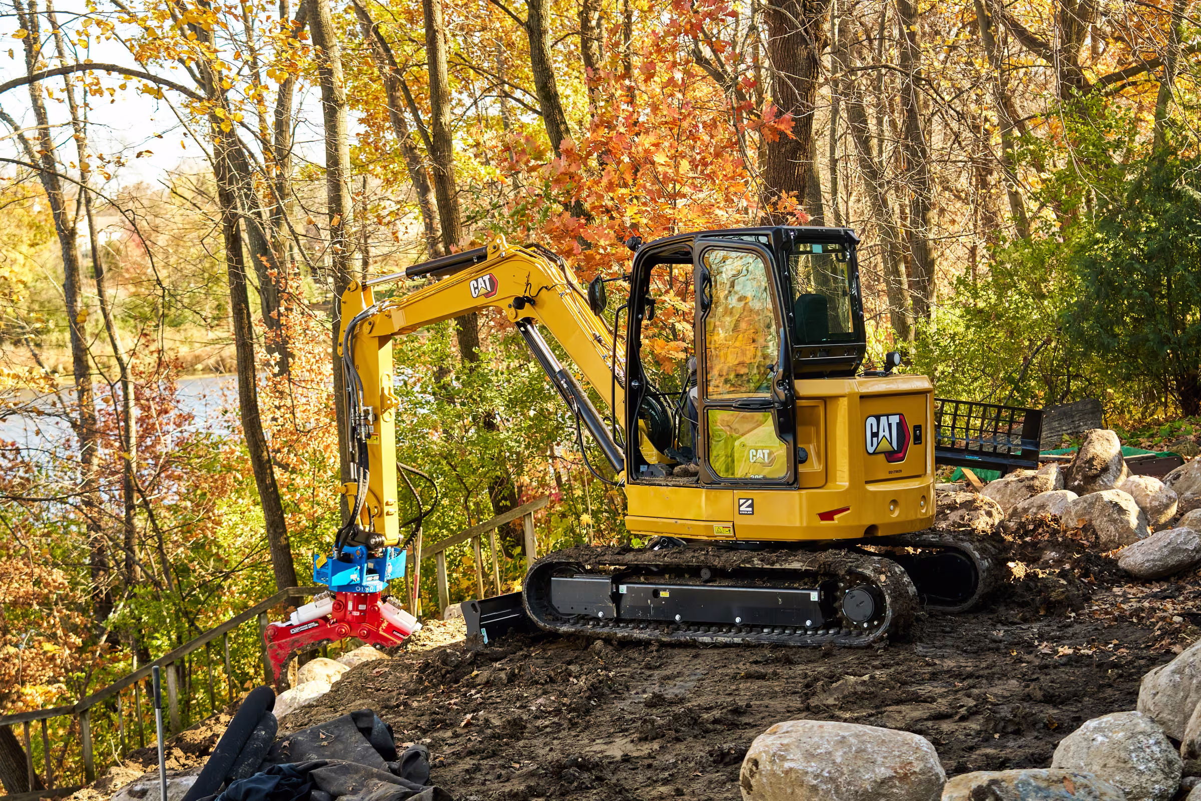 A yellow excavator sits among boulders on a wooded slope in Plymouth during autumn