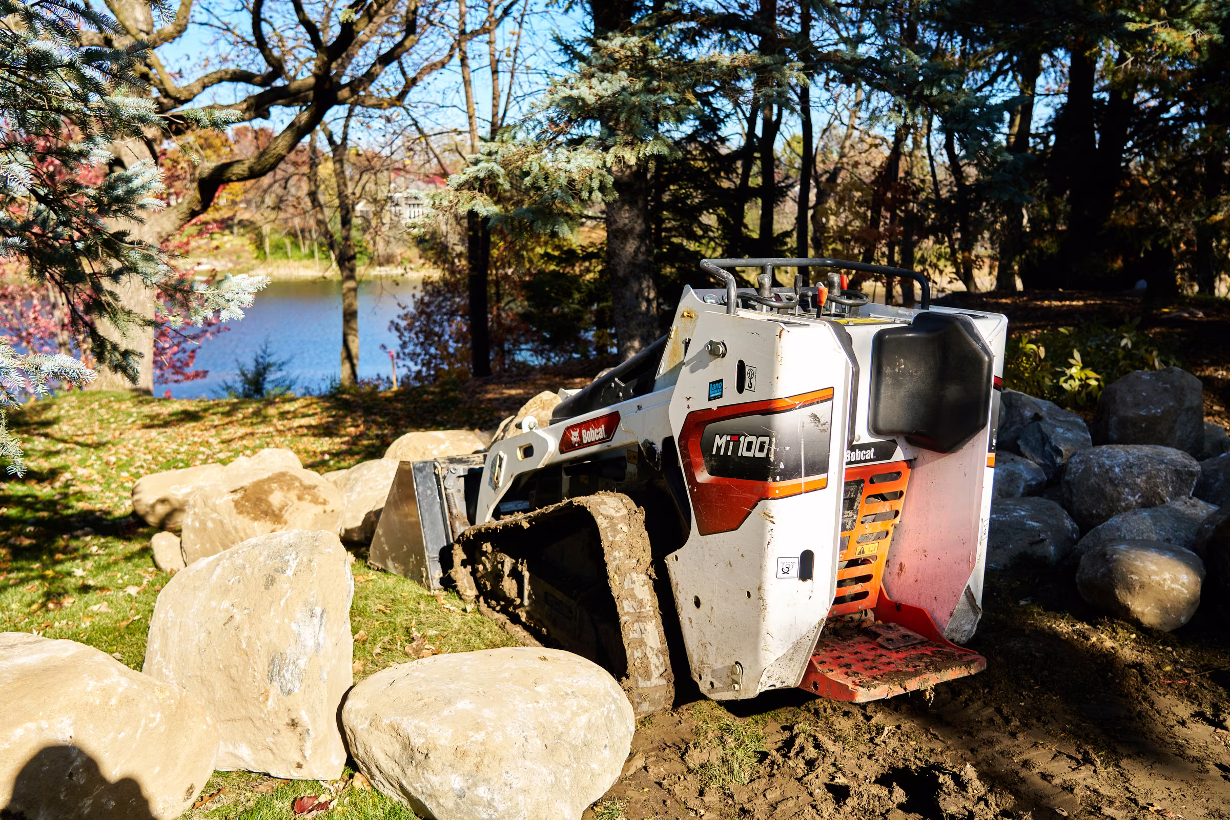 A compact tracked loader stands among large boulders with autumn trees and water beyond the work site
