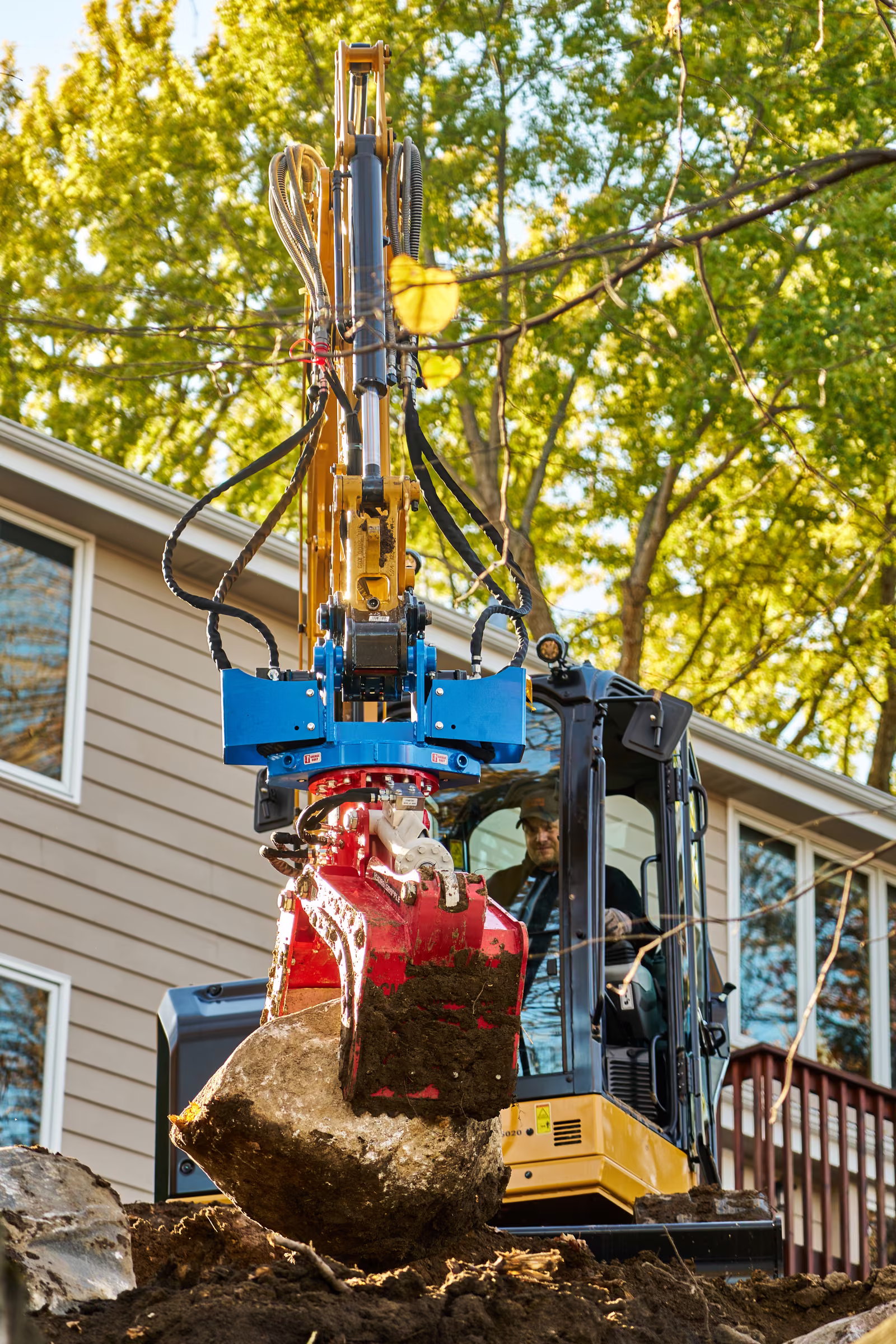 The excavator’s red hydraulic grapple holds a large boulder with the operator visible in the cab