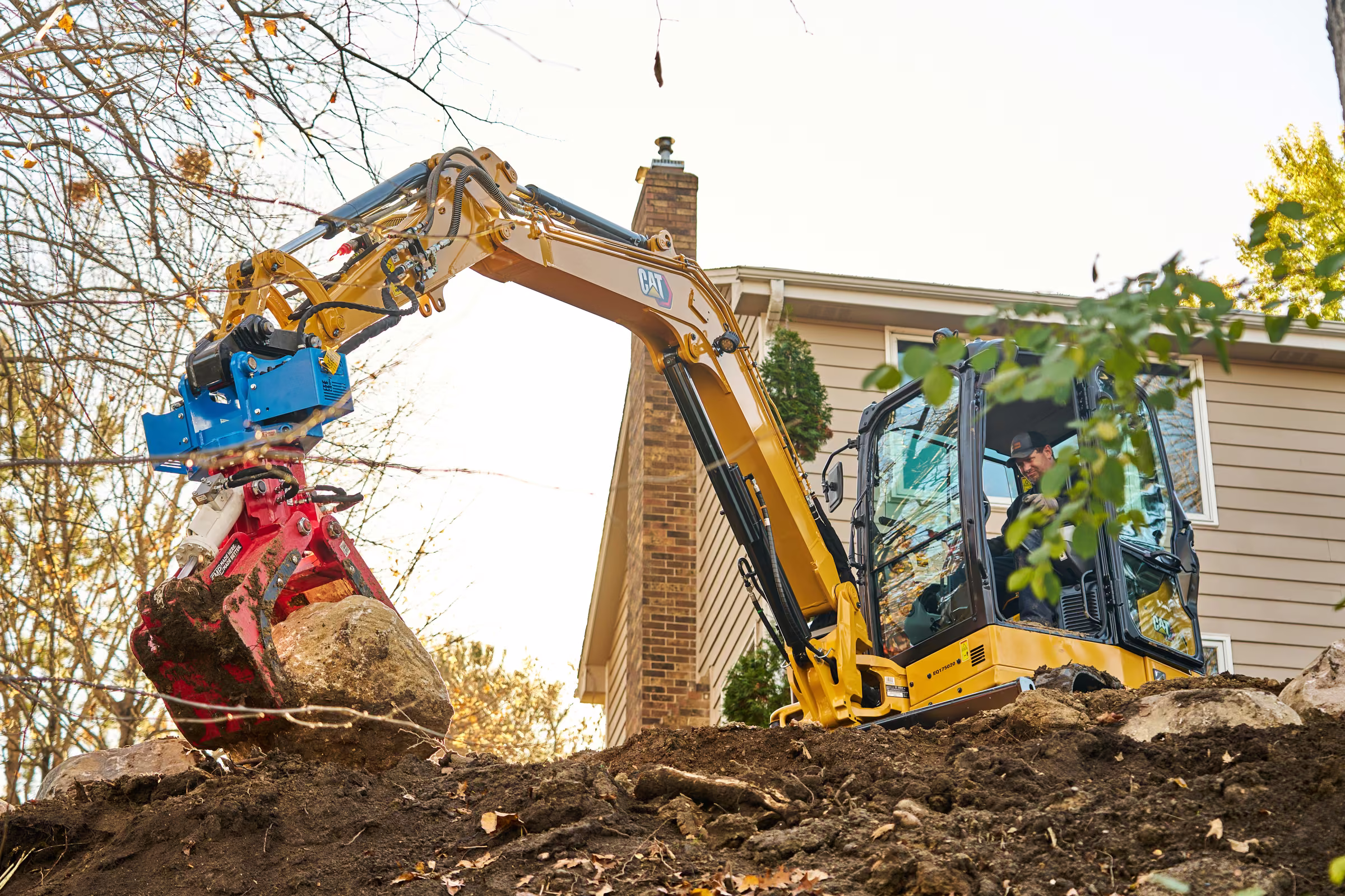 A yellow excavator carries a large boulder in a red hydraulic grapple beside a Plymouth home
