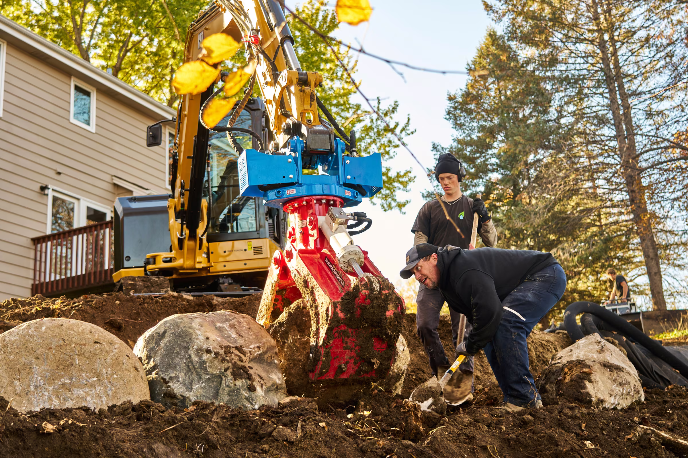 Two Freshwater Landscaping crew members guide work around a large boulder beneath the excavator’s grapple