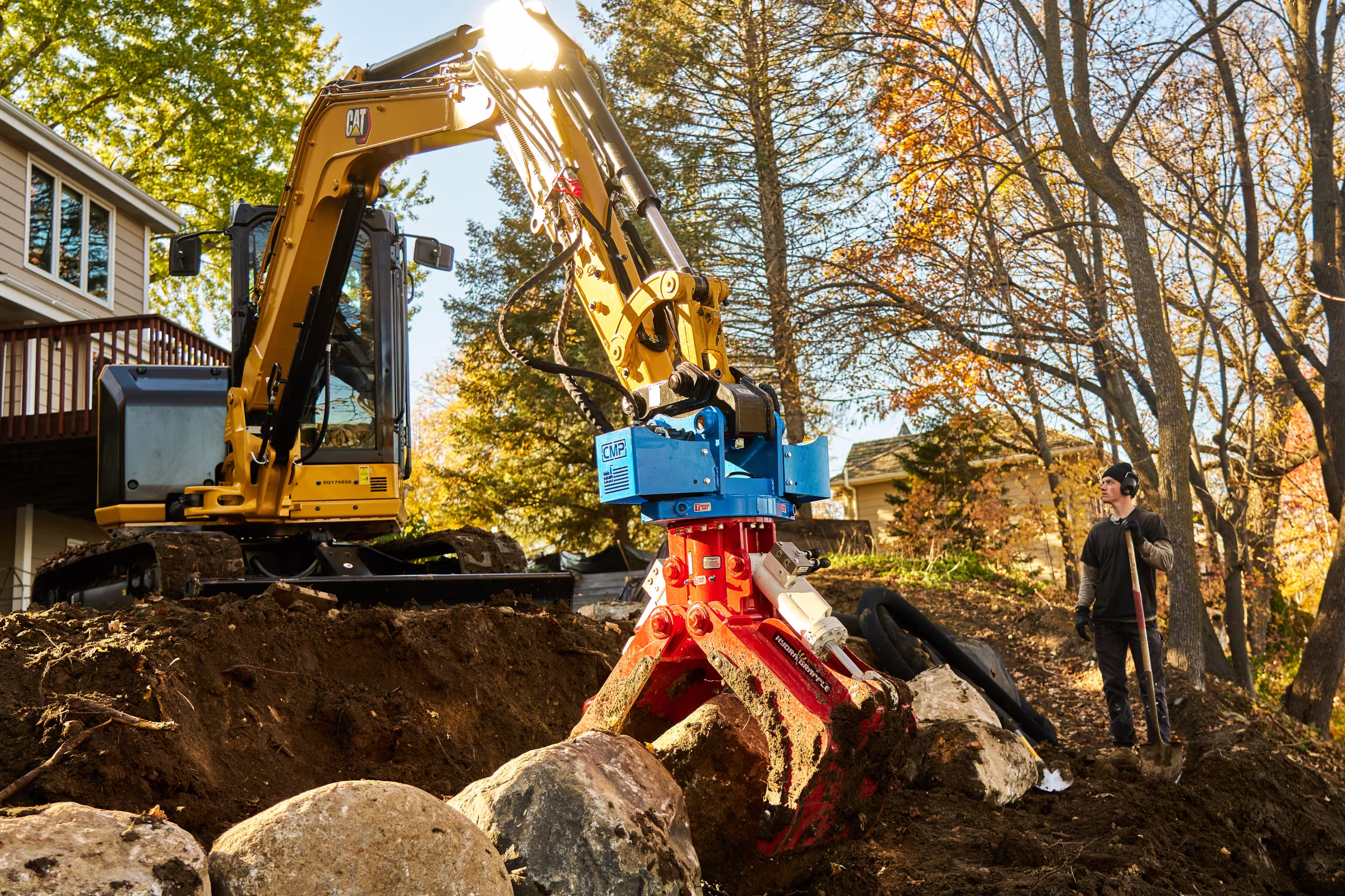 A crew member stands beside the excavator as its red grapple holds a boulder among stones at the work site