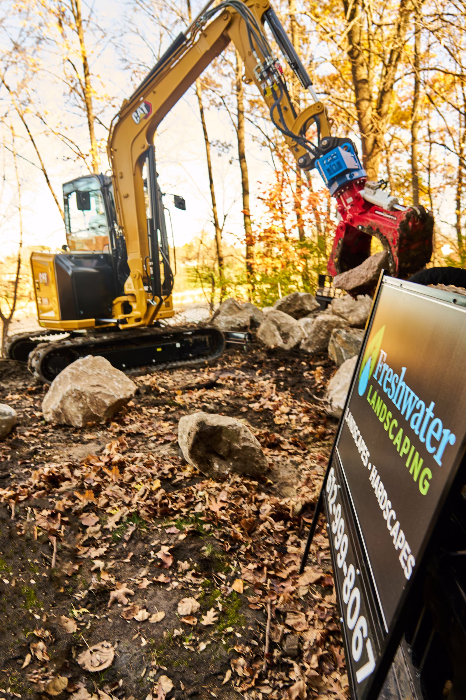 Freshwater Landscaping’s sign stands in the foreground while an excavator handles a boulder behind it