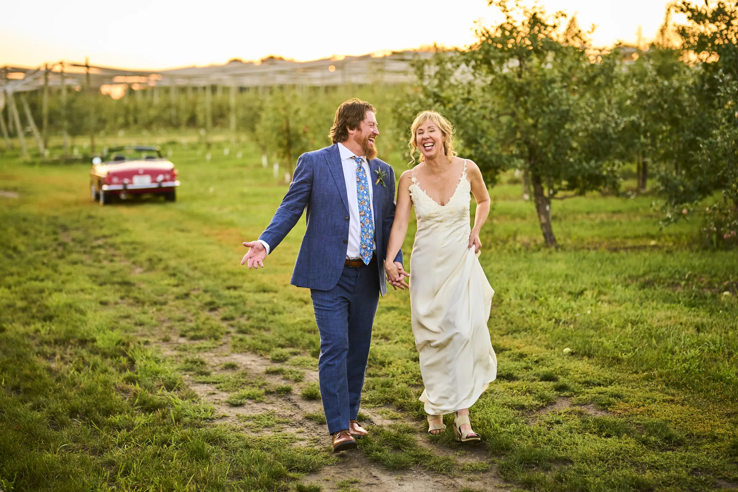Jeanne and Brian walk smiling through the orchard with their red convertible behind them in warm sunset light, Ashery Lane Farm, Mayer, Minnesota