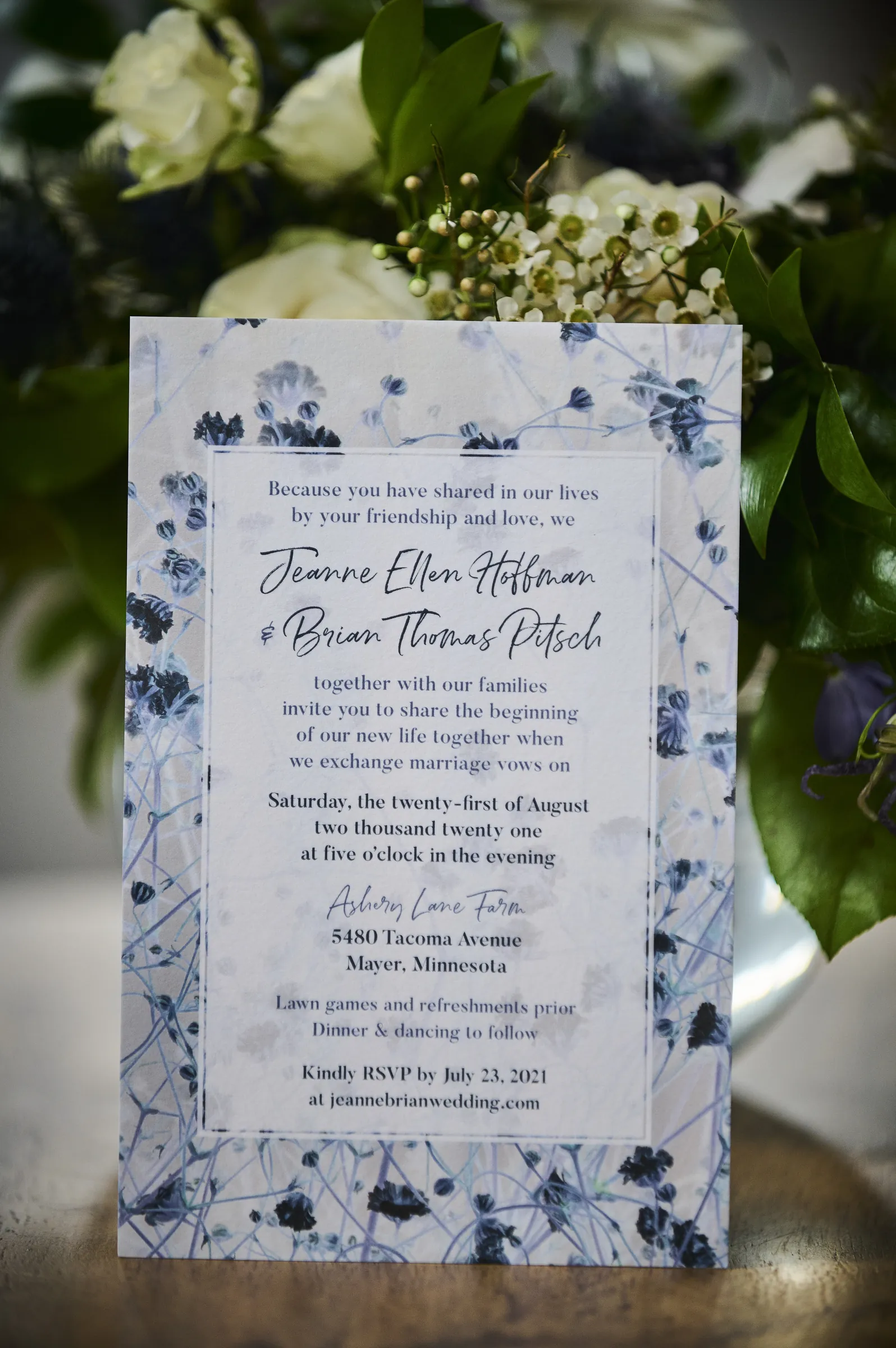 A close view of Jeanne Ellen Hoffman and Brian Thomas Pitsch’s wedding invitation surrounded by white flowers and green leaves, Ashery Lane Farm, Mayer, Minnesota