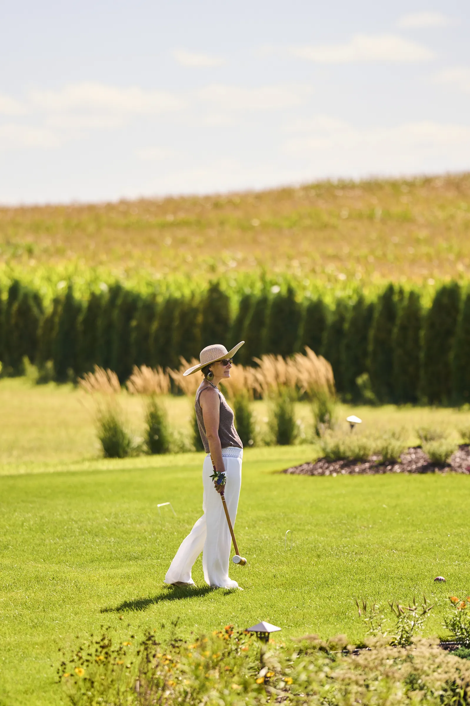 A guest in a broad-brimmed hat walks across the sunny lawn with fields and tall hedges beyond, Ashery Lane Farm, Mayer, Minnesota