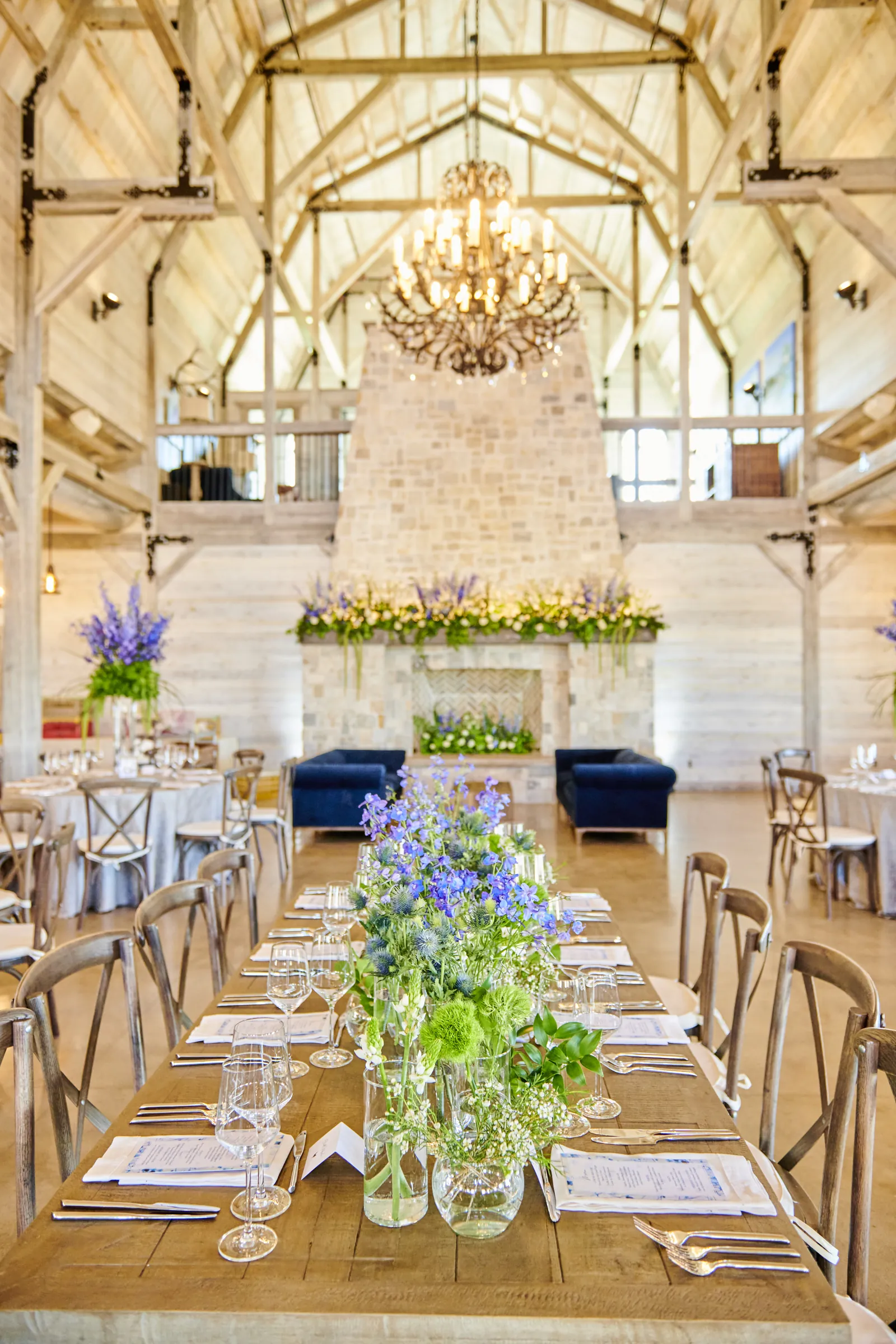 A long wooden reception table with blue and white flowers beneath a chandelier and exposed barn beams, Ashery Lane Farm, Mayer, Minnesota