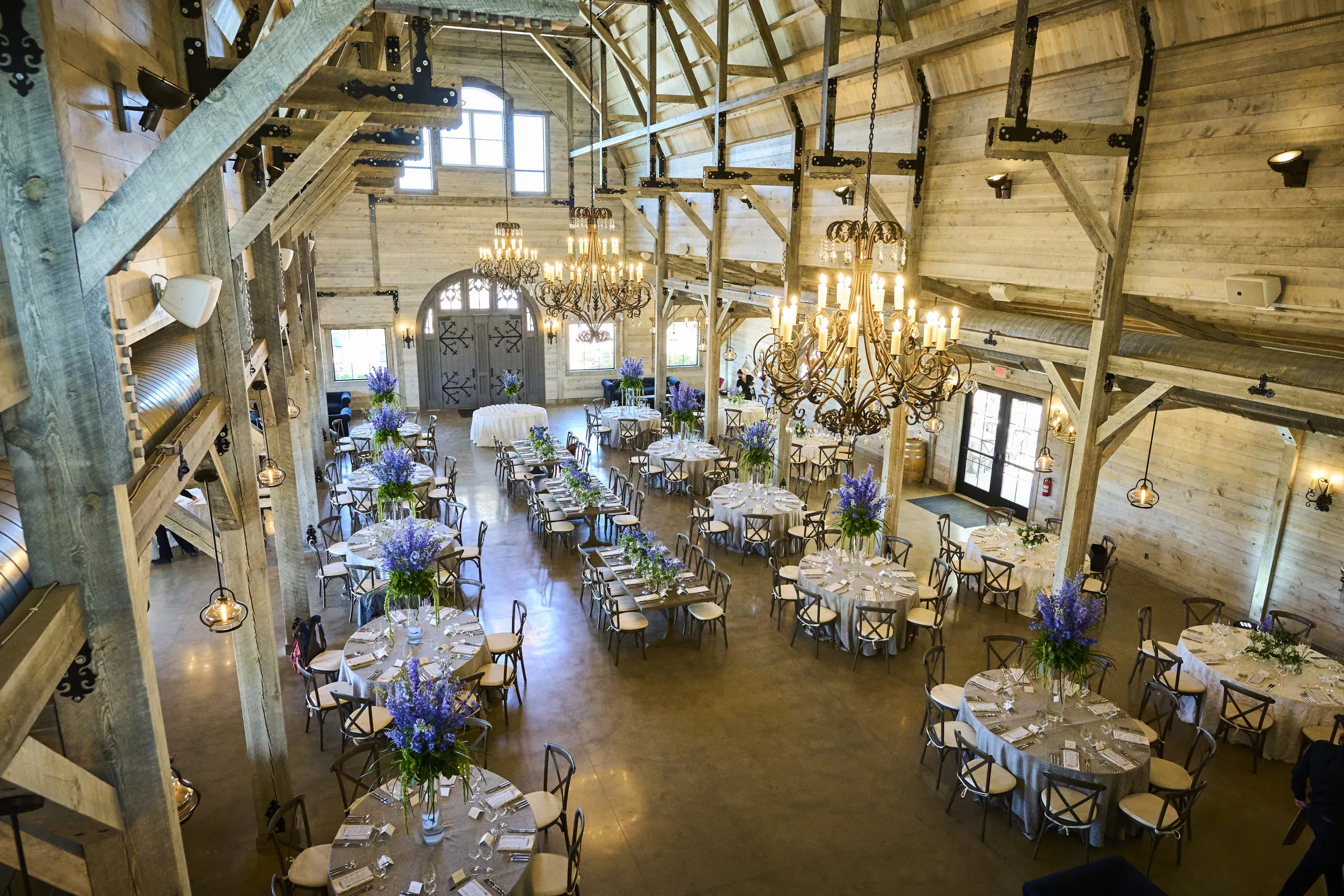 An elevated view of wooden tables, flowers, chandeliers, and exposed beams inside the reception barn, Ashery Lane Farm, Mayer, Minnesota