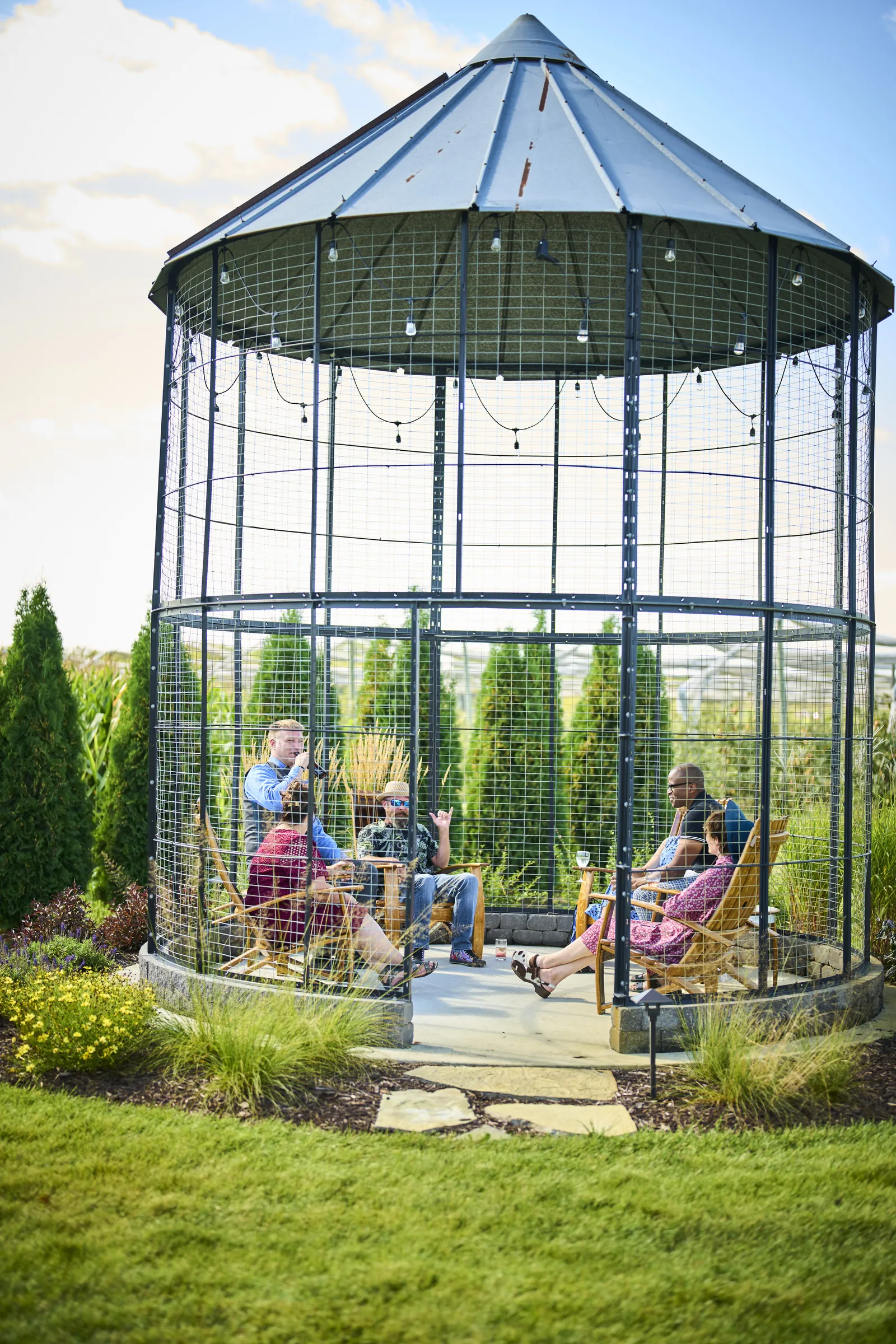 Wedding guests sit together inside an open metal gazebo in the gardens, Ashery Lane Farm, Mayer, Minnesota