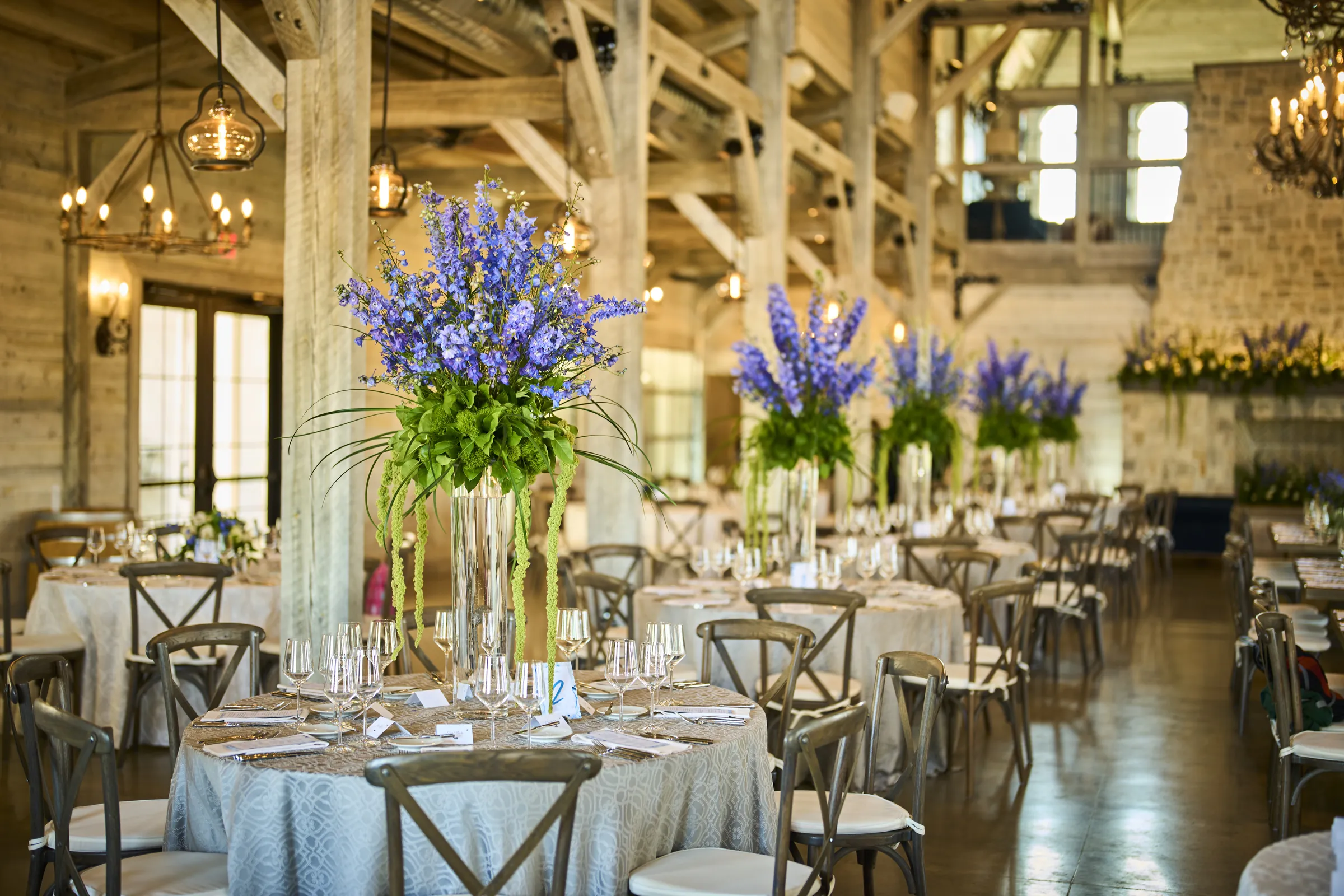 Tall blue and purple flowers rise above a wooden reception table with glassware and candles, Ashery Lane Farm, Mayer, Minnesota