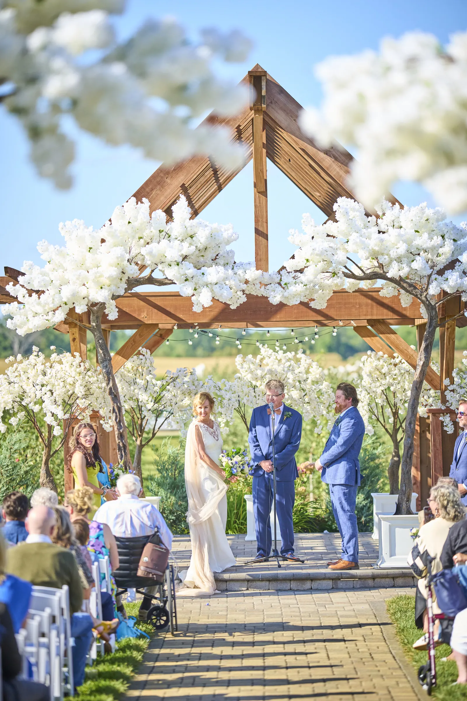 Jeanne and Brian stand with their officiant beneath a wooden arbor covered in white blossoms, Ashery Lane Farm, Mayer, Minnesota