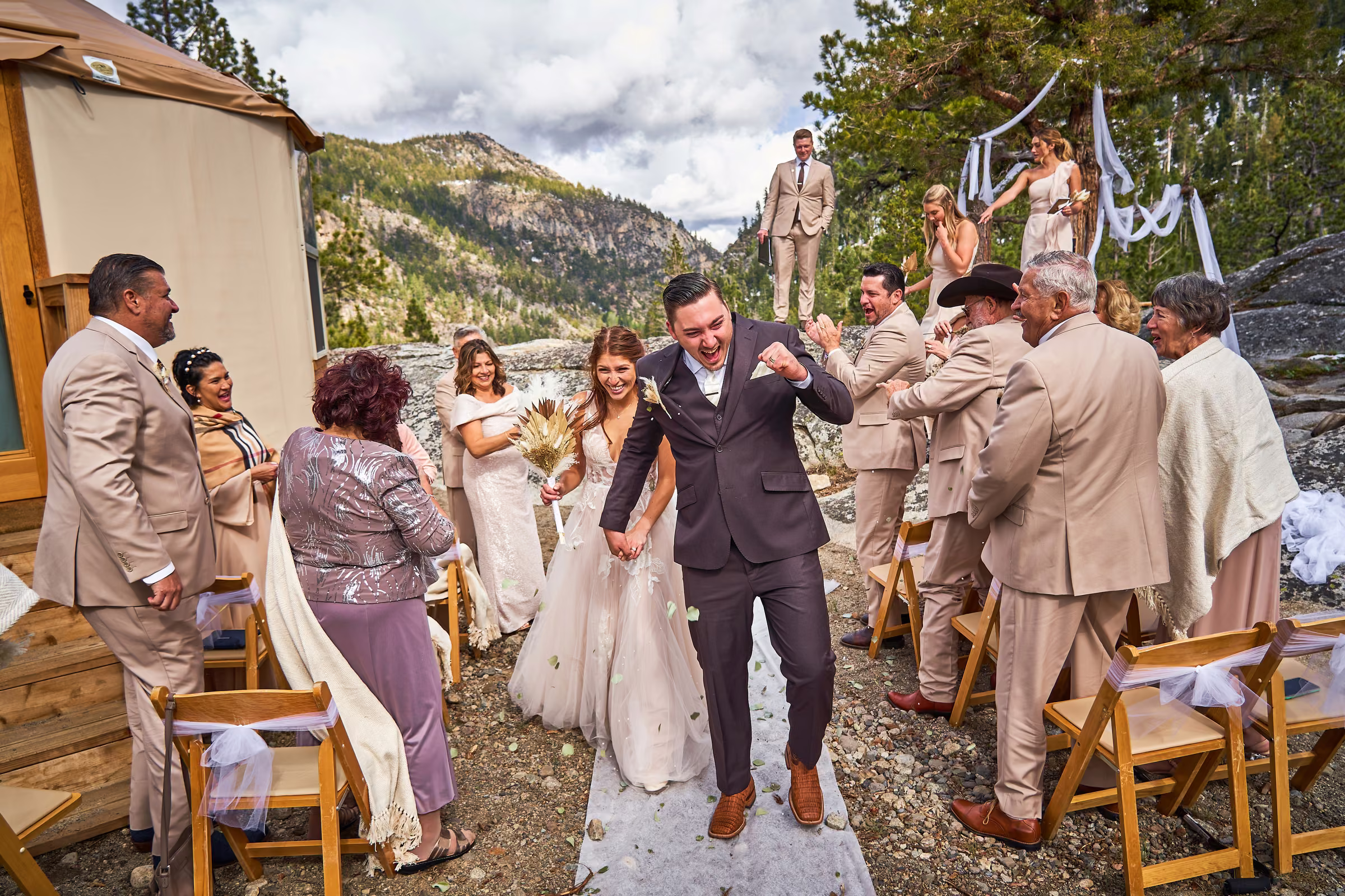 Joe and Gabby smile and raise their hands as they walk back down the aisle between their guests, Hope Valley, California