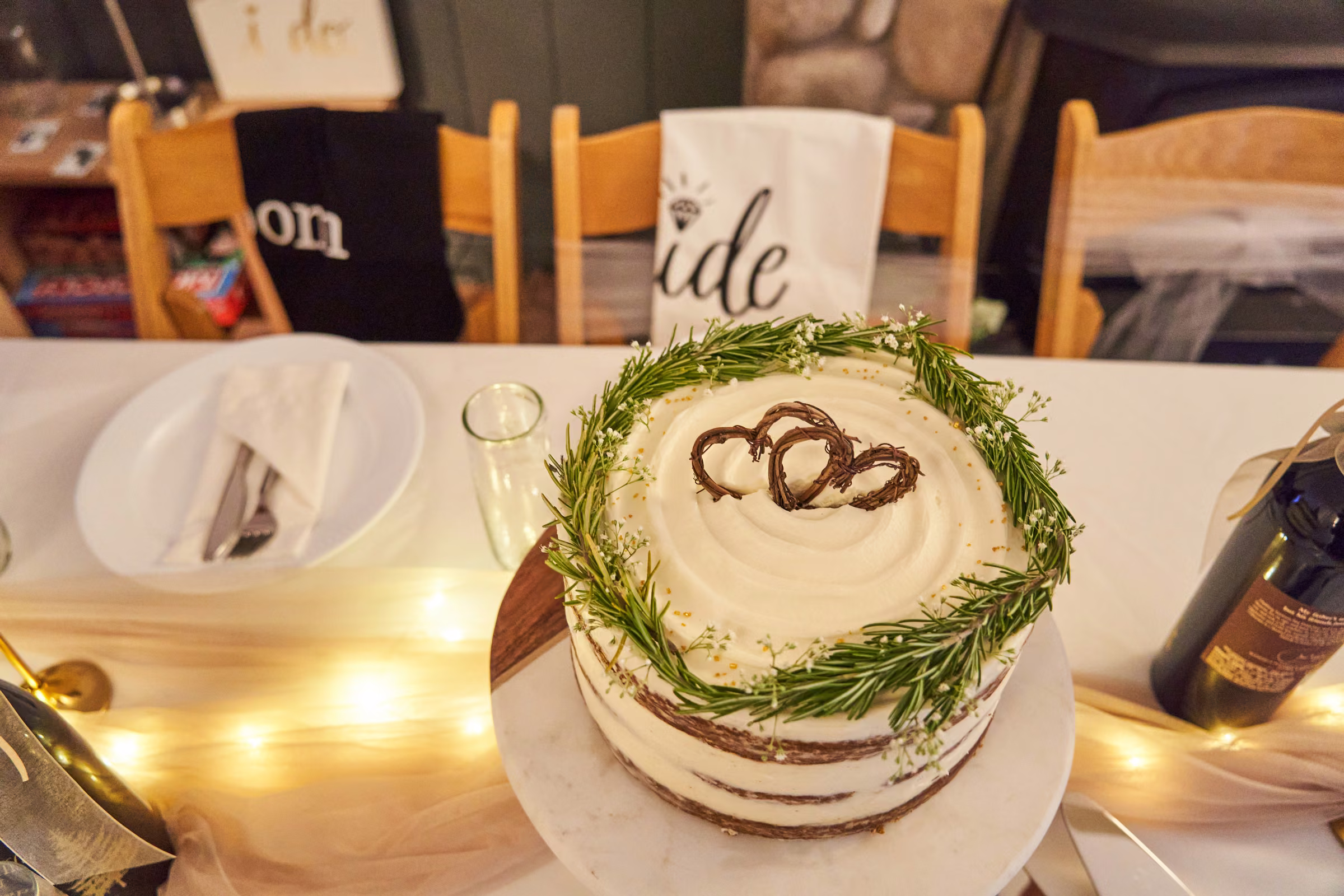 A small wedding cake decorated with greenery and two intertwined heart shapes sits on the reception table, Hope Valley, California