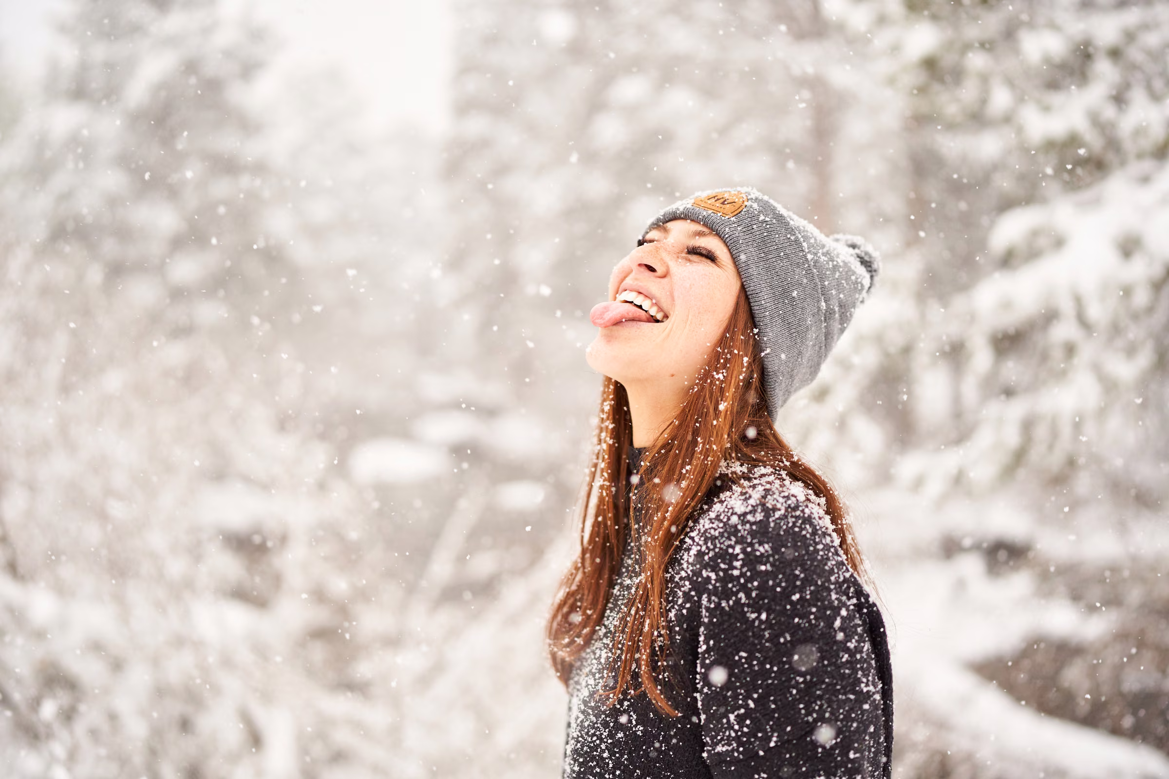 Gabby smiles upward in a knit hat as snow falls around her among the trees, Hope Valley, California