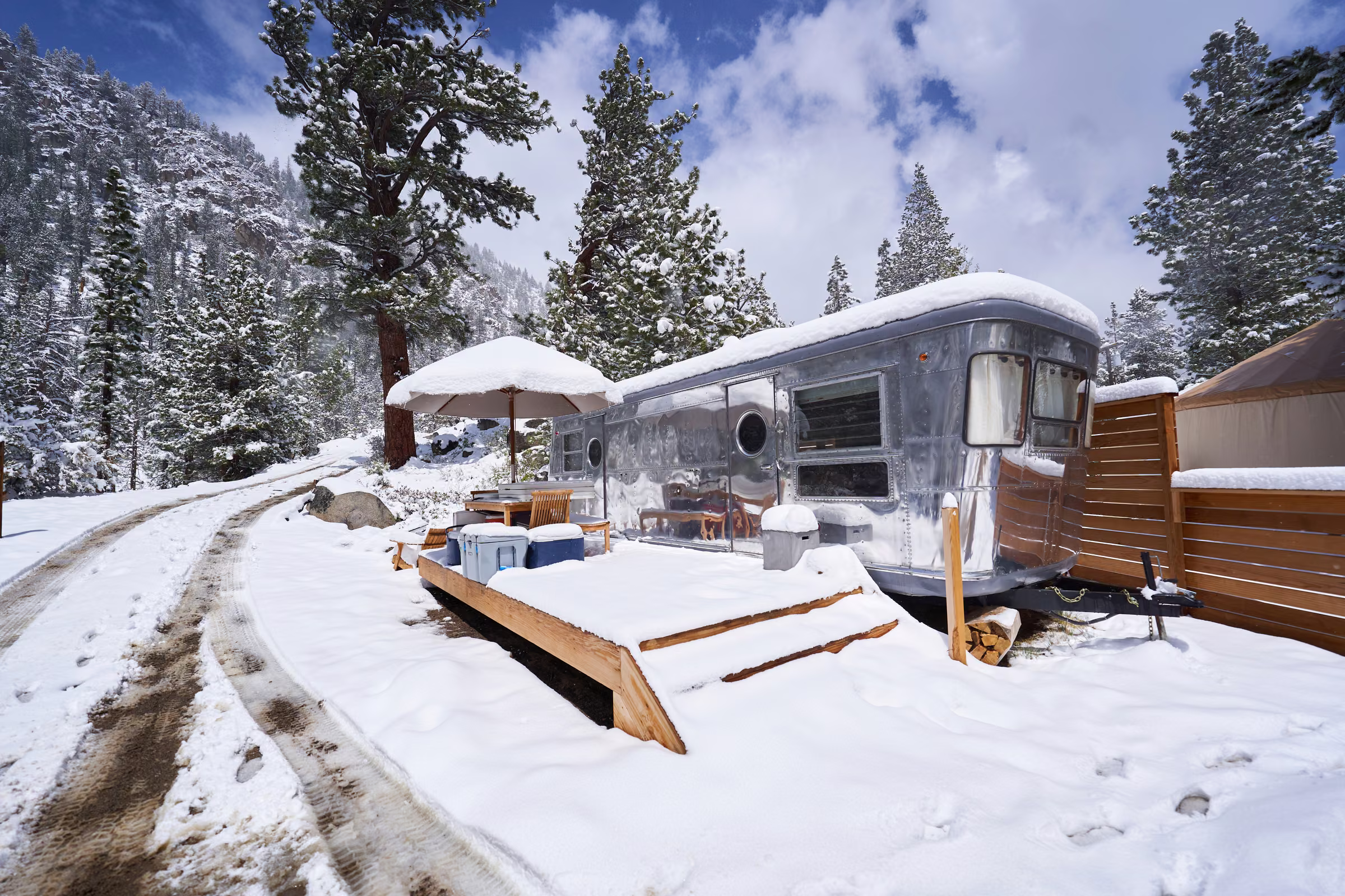 A silver travel trailer and its wooden deck sit beside a snow-covered road among tall pine trees, Hope Valley, California
