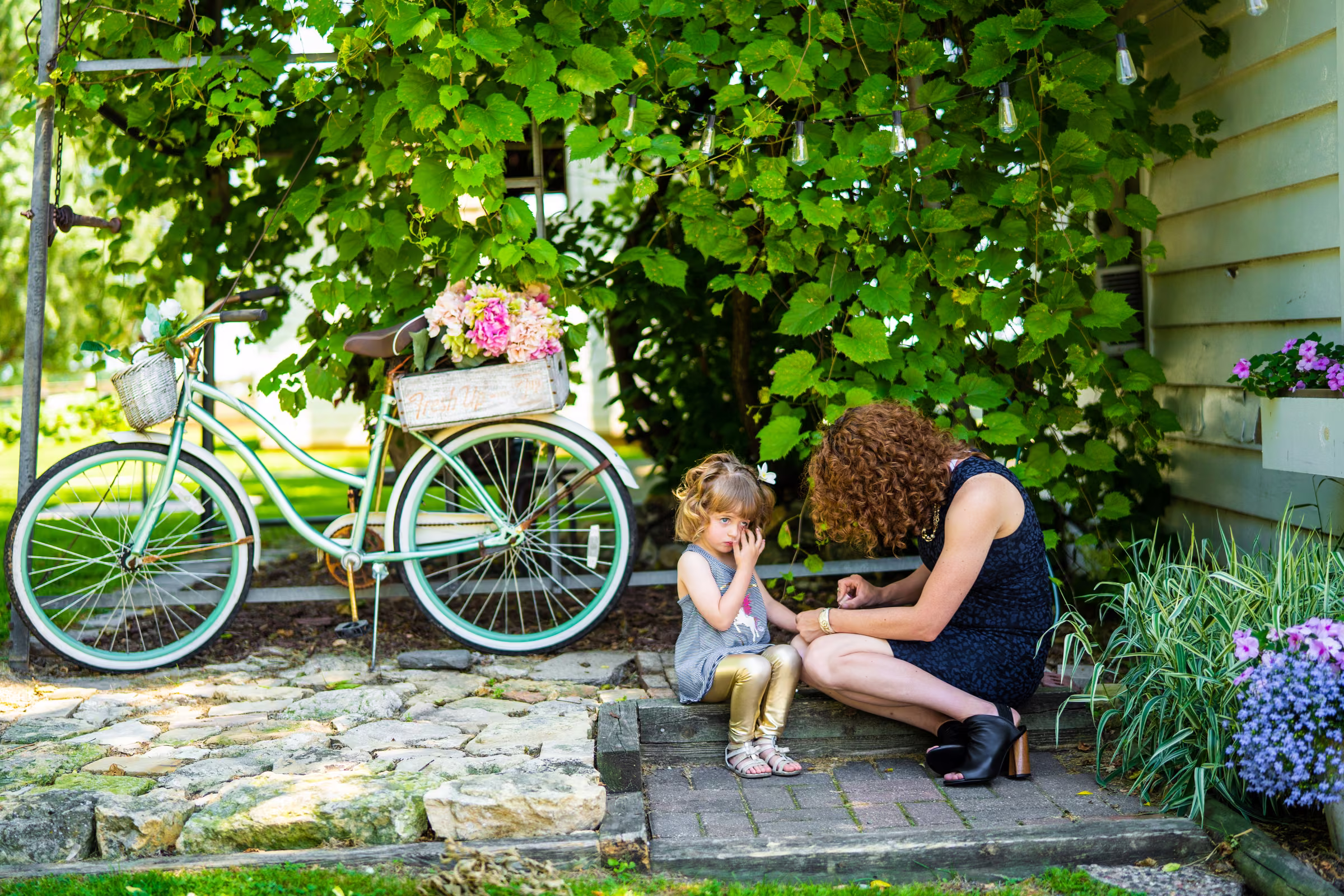 A mother paints a flower girl’s nails beside a green bicycle decorated with flowers